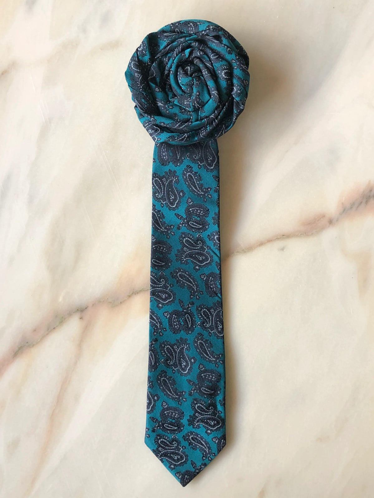 Broochella Fuente Rosette Tie Brooch - Maurizio in teal with a dark grey paisley pattern, finished with a sculptural rosette and photographed on a white marble background.