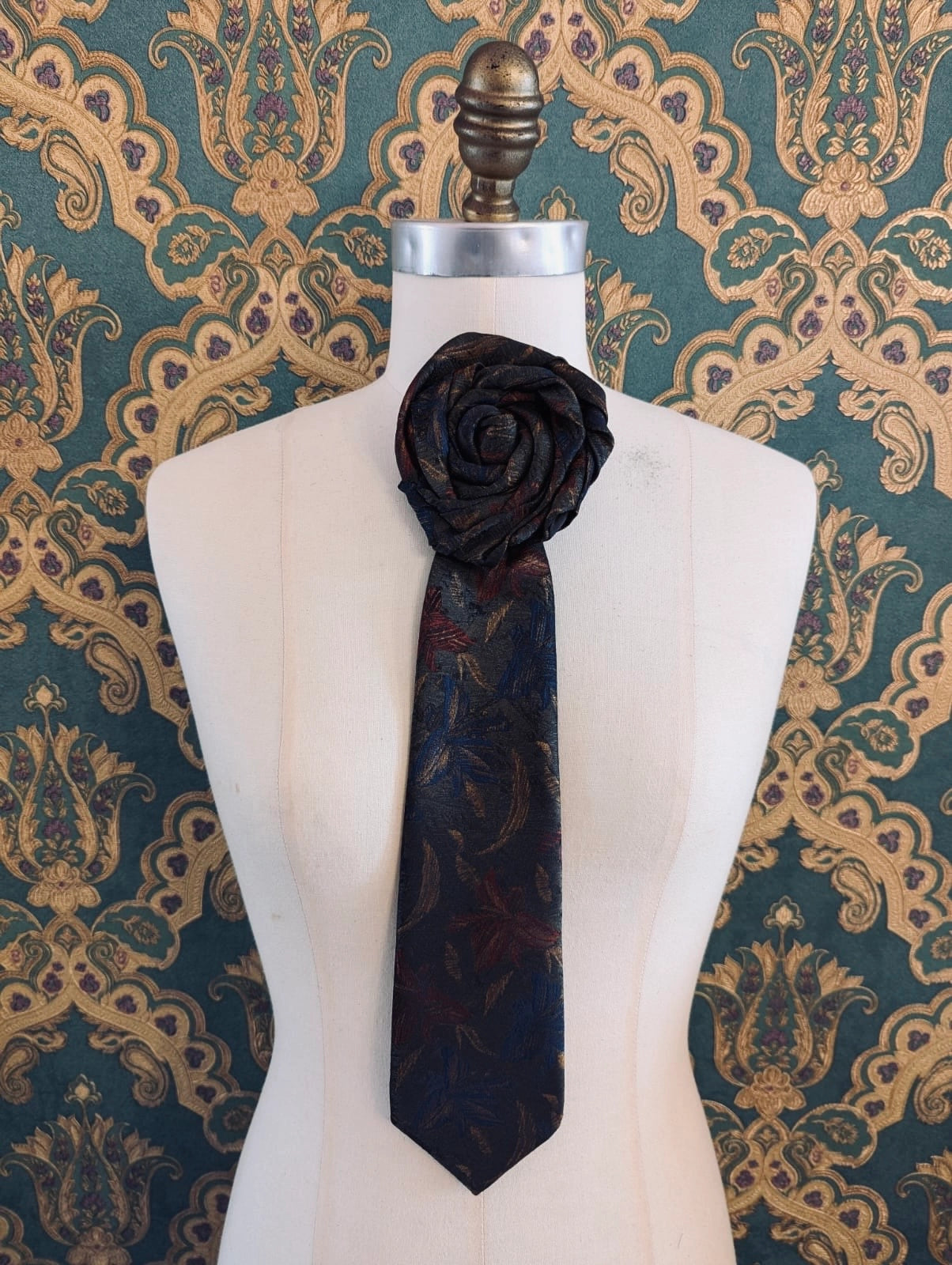 Broochella Fuente Ignazio vintage up-cycled vintage handcrafted rosette tie in blue and green. Pinned on a mannequin. The background is green and gold damask wallpaper