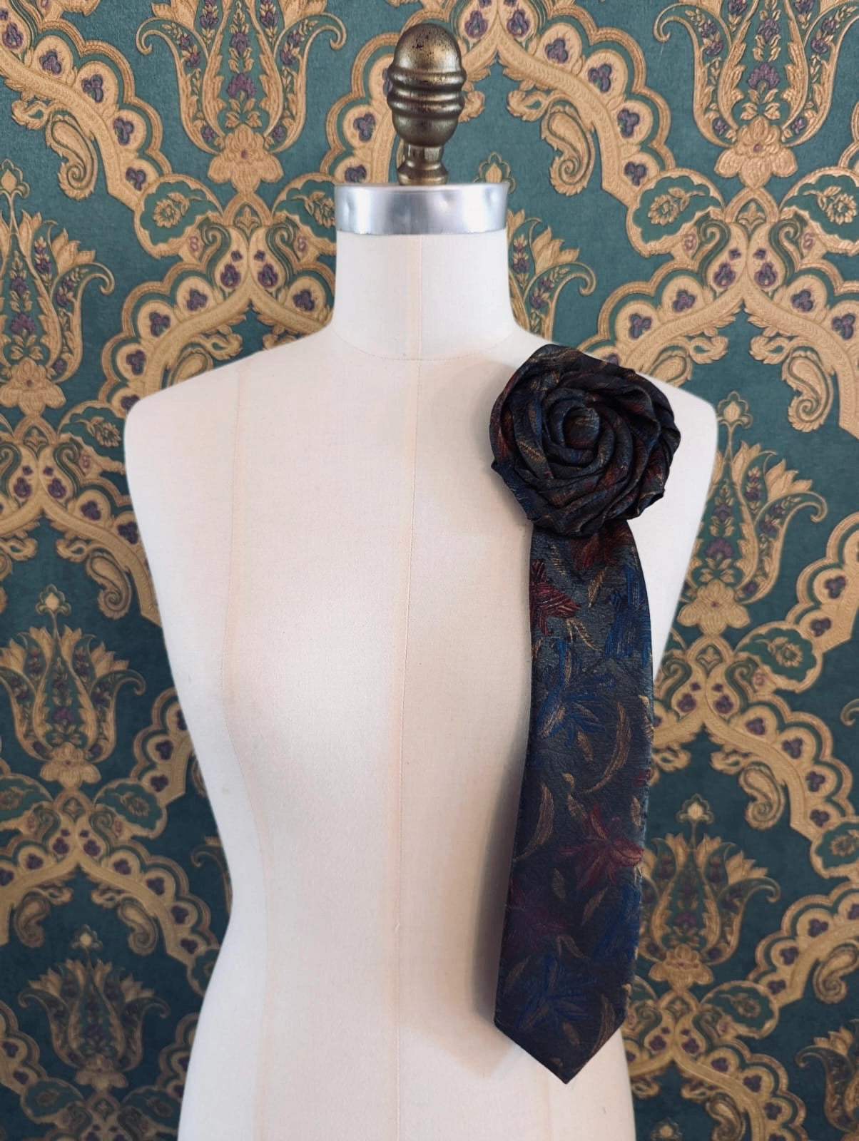 Broochella Fuente Ignazio vintage up-cycled vintage handcrafted rosette tie in blue and green. Pinned on a mannequin. The background is green and gold damask wallpaper