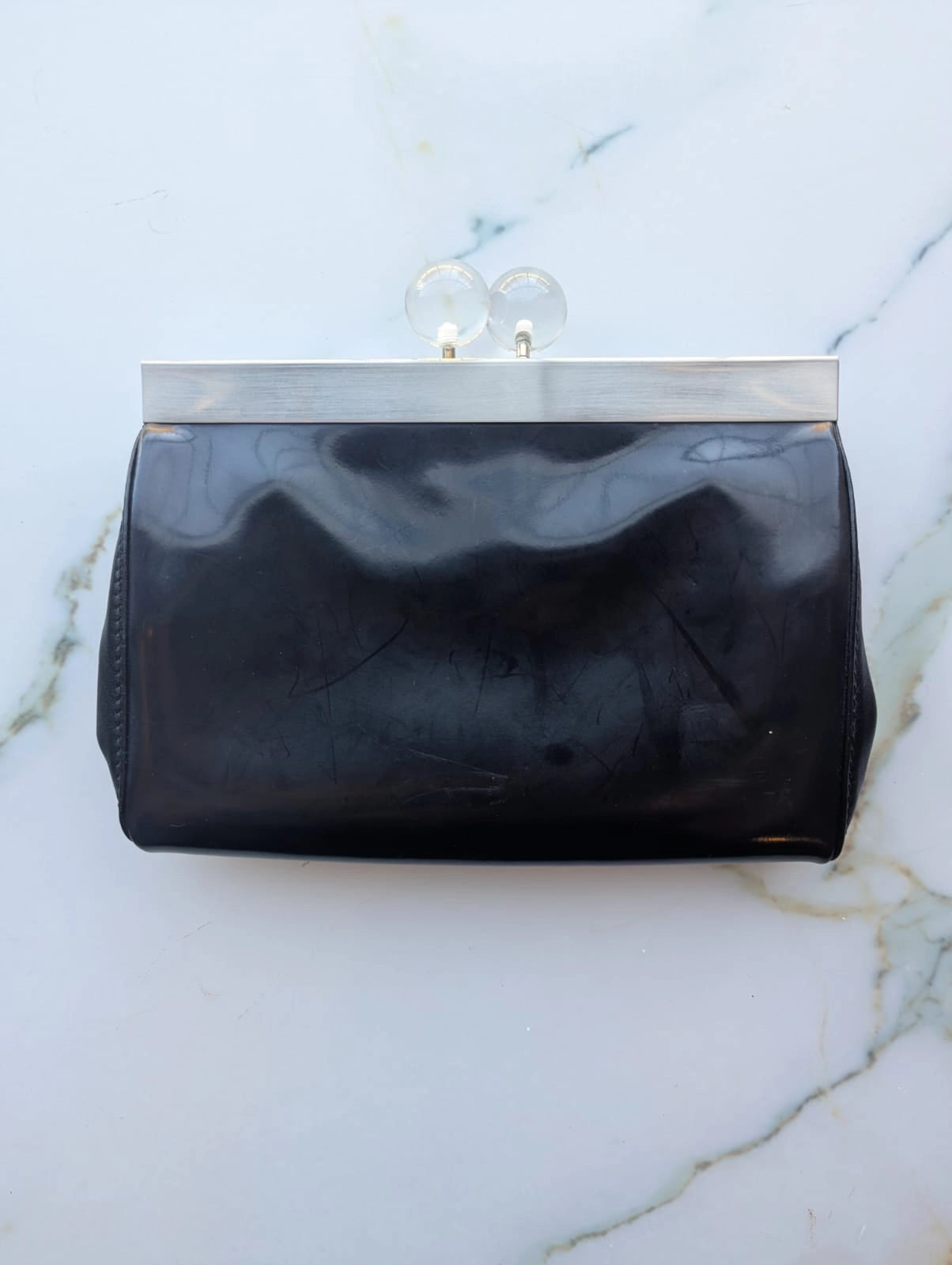 A Broochella The Stylist's Closet Furla black leather clutch purse with a silver-tone metal frame and clear acrylic clasp, displayed flat on a white marble surface.