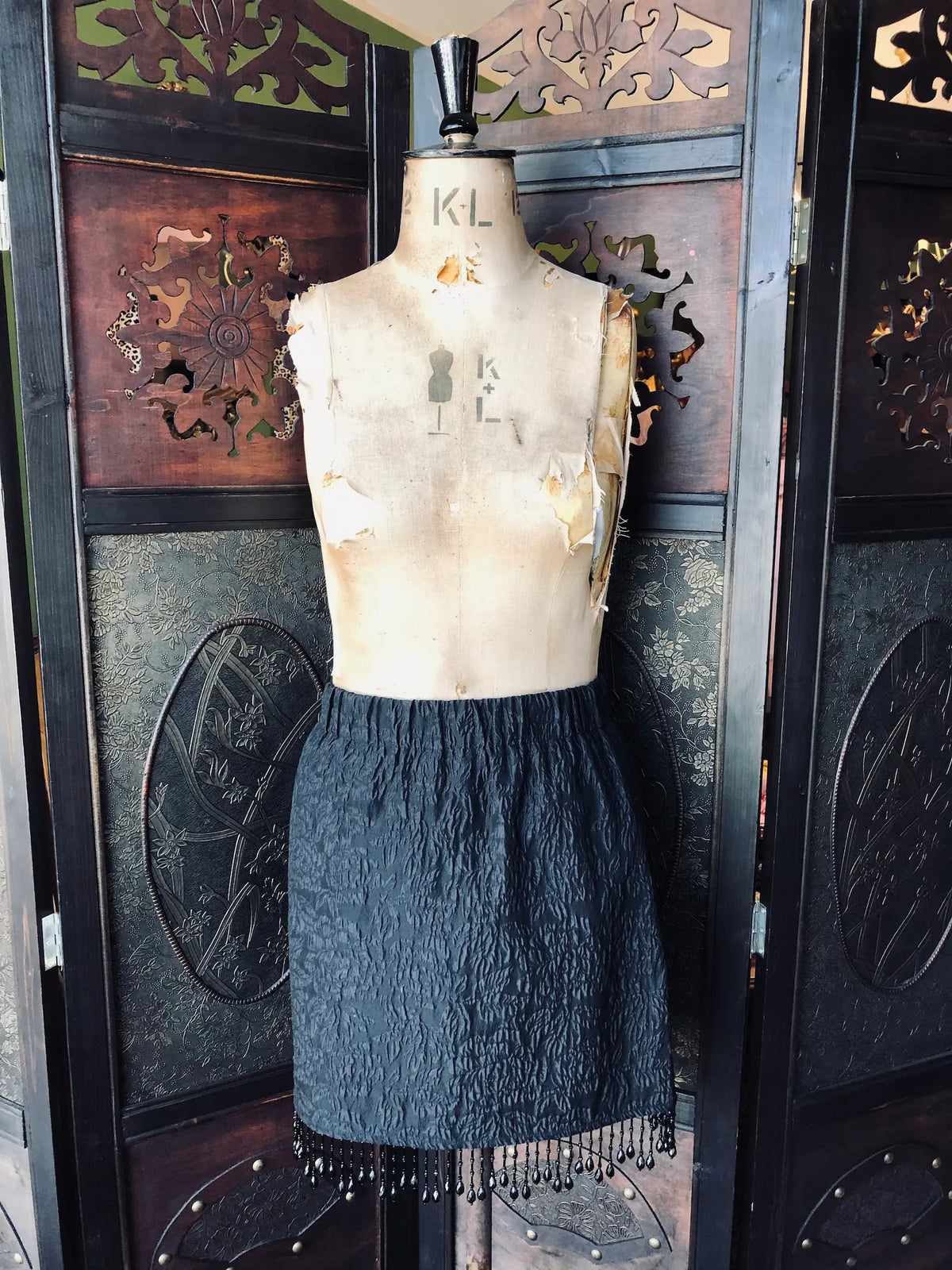 Broochella Mannequin wearing a Ganni black textured skirt with fringe against an ornate wooden background