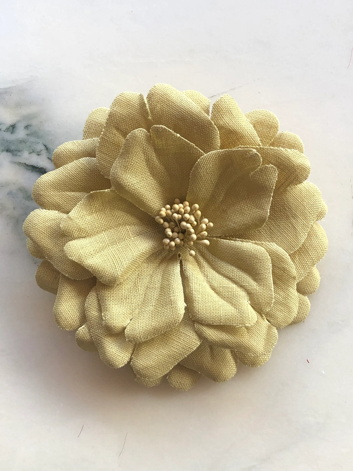 Broochella Men's Garofano Flower Brooch in beige, featuring full layered fabric petals with a textured stamen centre, photographed on a cream marble background.