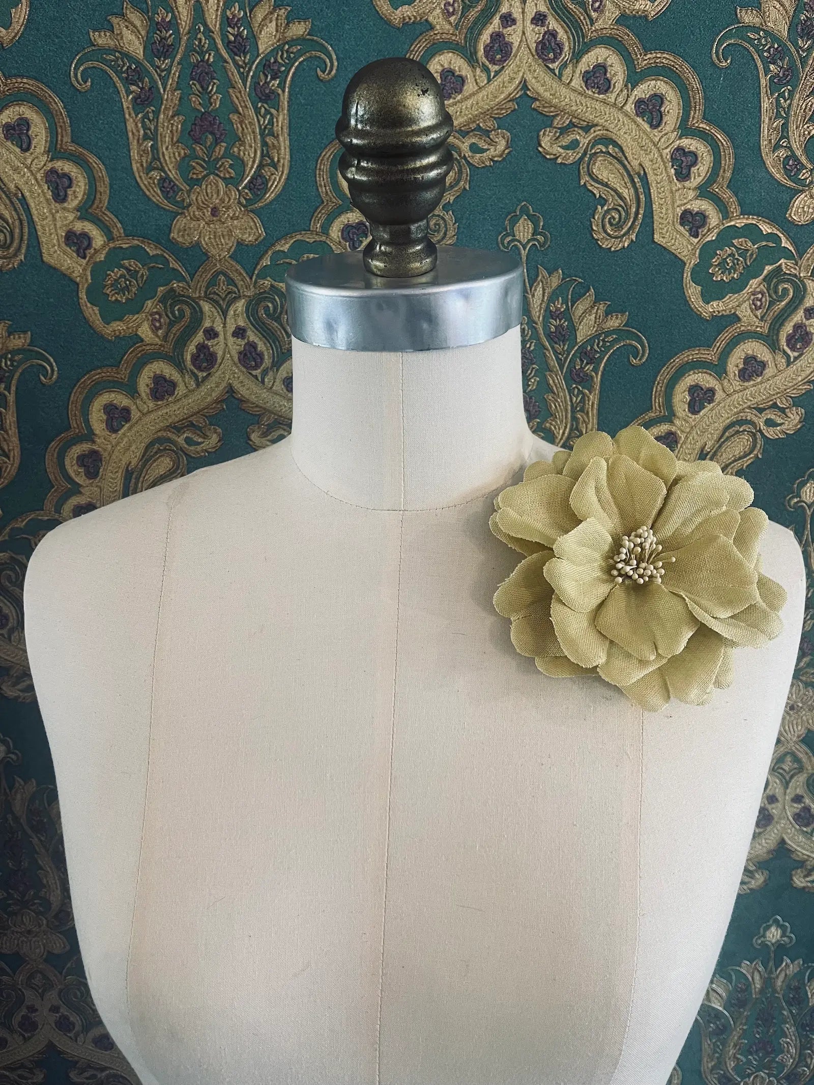 Broochella garofano handmade fabric flower brooch in beige. Styled on a mannequin. The background is green and gold damask wallpaper