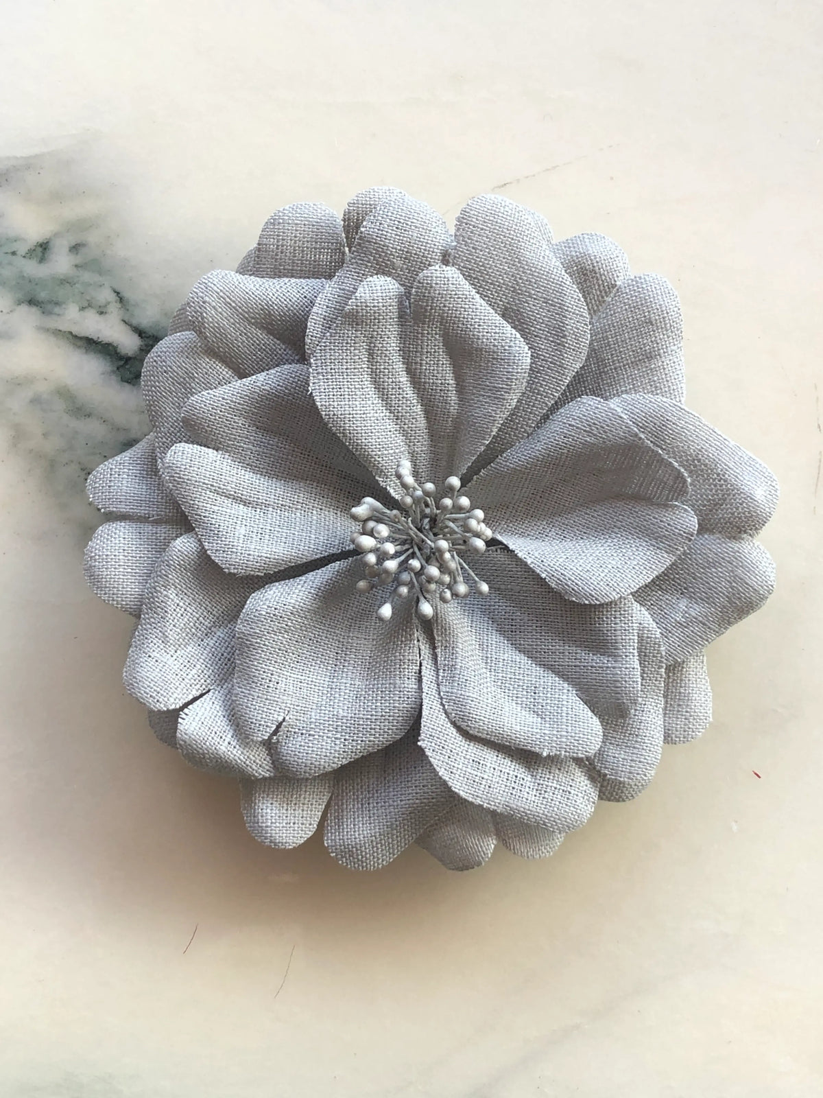 Broochella Men's Garofano Flower Brooch in soft grey, featuring full layered fabric petals with a tonal textured stamen centre, photographed on a cream marble background.