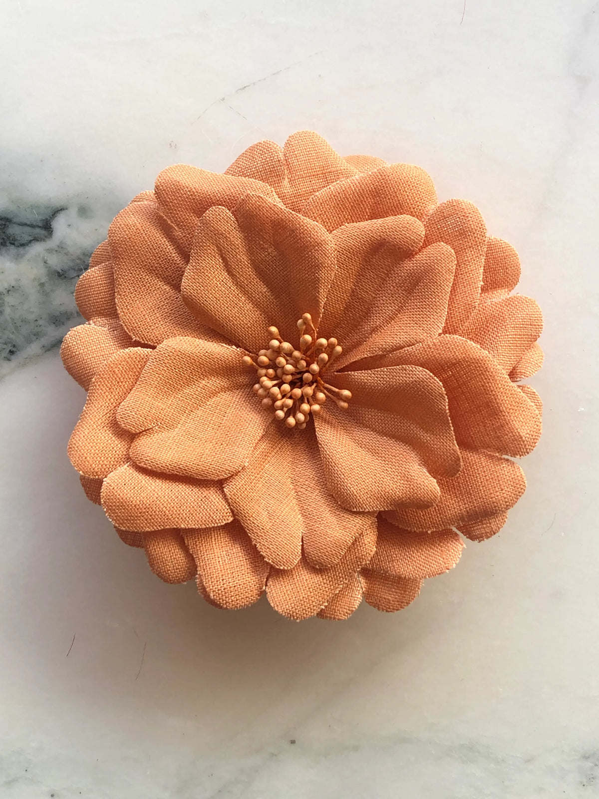 Broochella Men's  Garofano Flower Brooch in warm orange, featuring full layered fabric petals with a tonal textured stamen centre, photographed on a cream marble background.