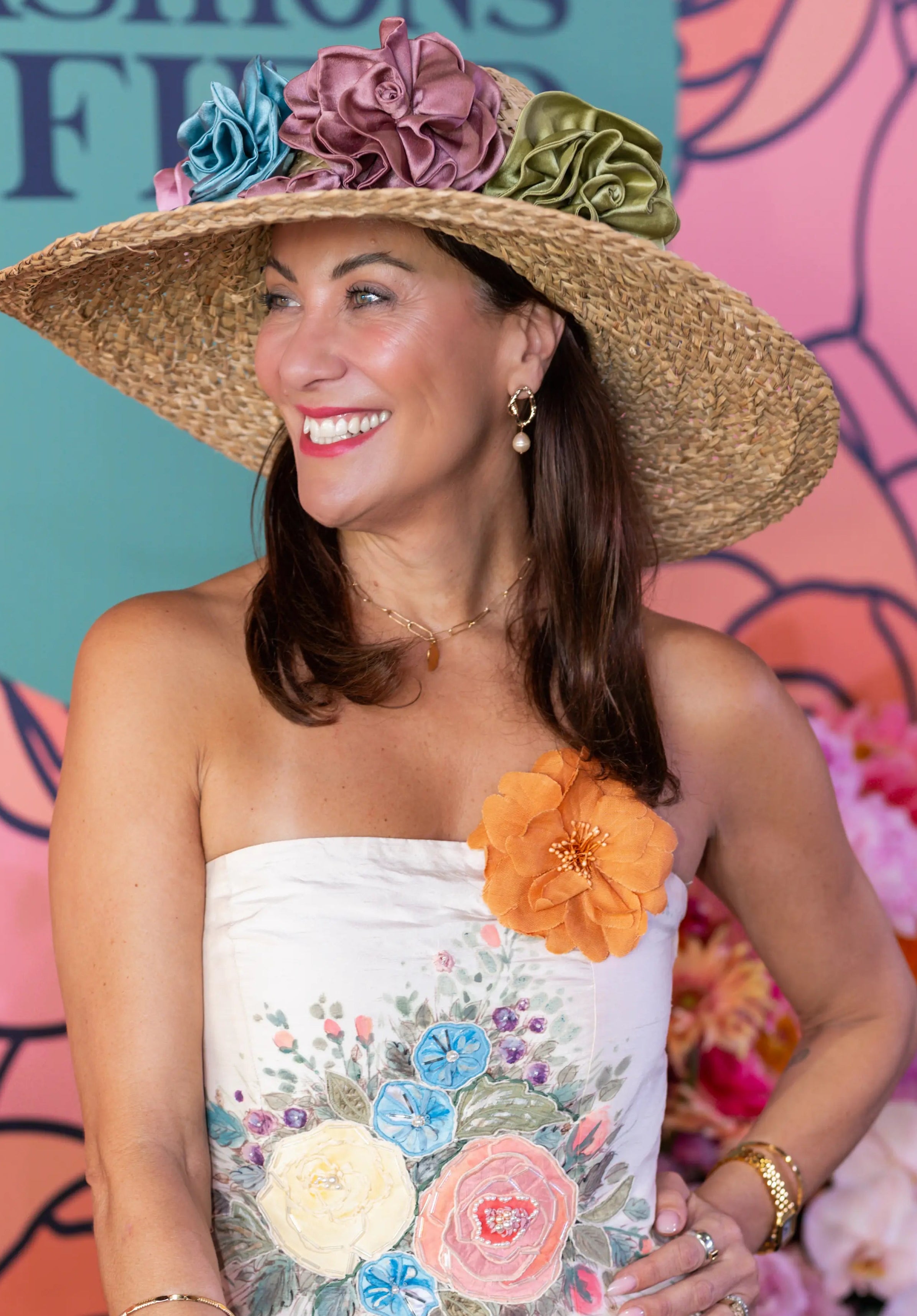 A woman wearing a strapless ivory  floral dress with a large straw hat adorned with fabric flowers, holding a tan clutch bag in front of a pink floral backdrop. SHe is wearing the Broochella garofano fabric flower brooch in orange. 