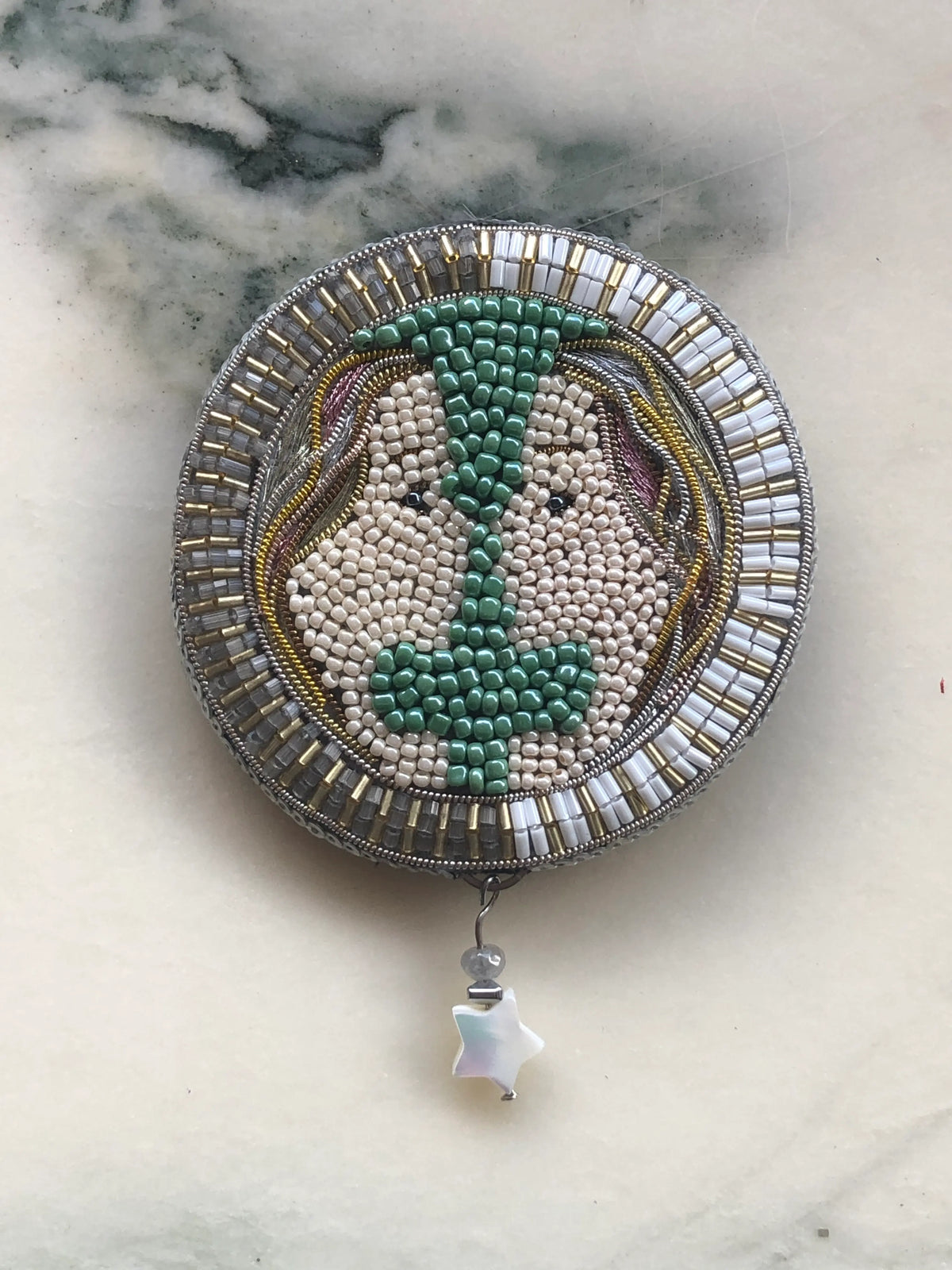 A Broochella Zodiac Gemini circular hand beaded brooch featuring a stylized face with green hair and a silver-toned beaded border, displayed on a light marble surface with a small dangling star charm.