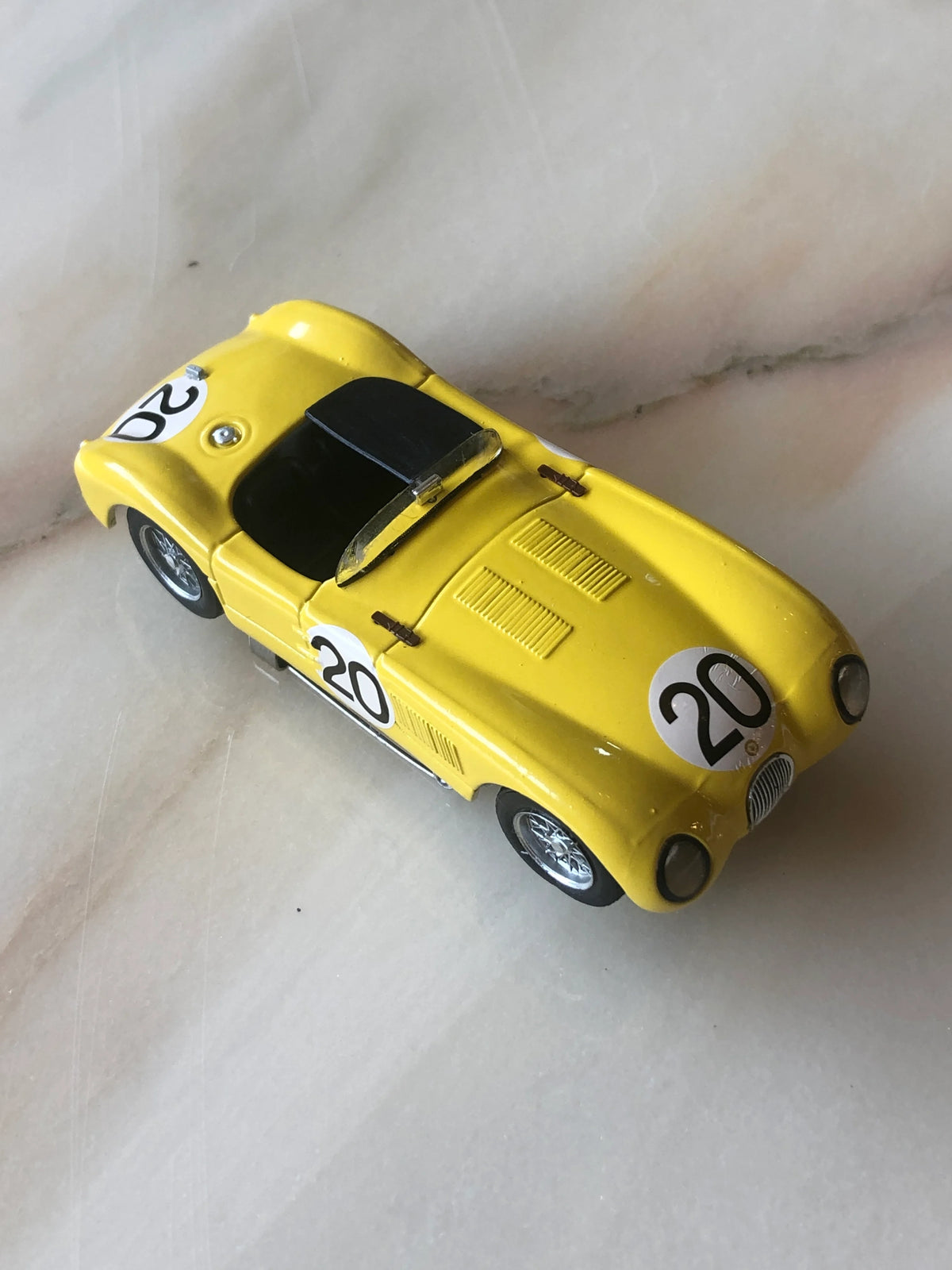 Broochella Giallo Corsa No.20 Vintage Racing Car Brooch photographed from an elevated front angle, featuring a bright yellow open-top sports car, black cockpit, number 20 racing decals, silver wire-spoke wheels, detailed bonnet vents and classic racing design.