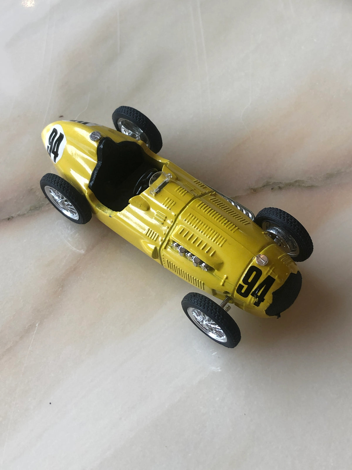 Broochella Giallo Corsa No.20 Vintage Racing Car Brooch shown from an elevated three-quarter angle, featuring a bright yellow open-cockpit racing car with black number 94 decals, silver wire-spoke wheels, exposed engine details, vented bonnet and black interior.