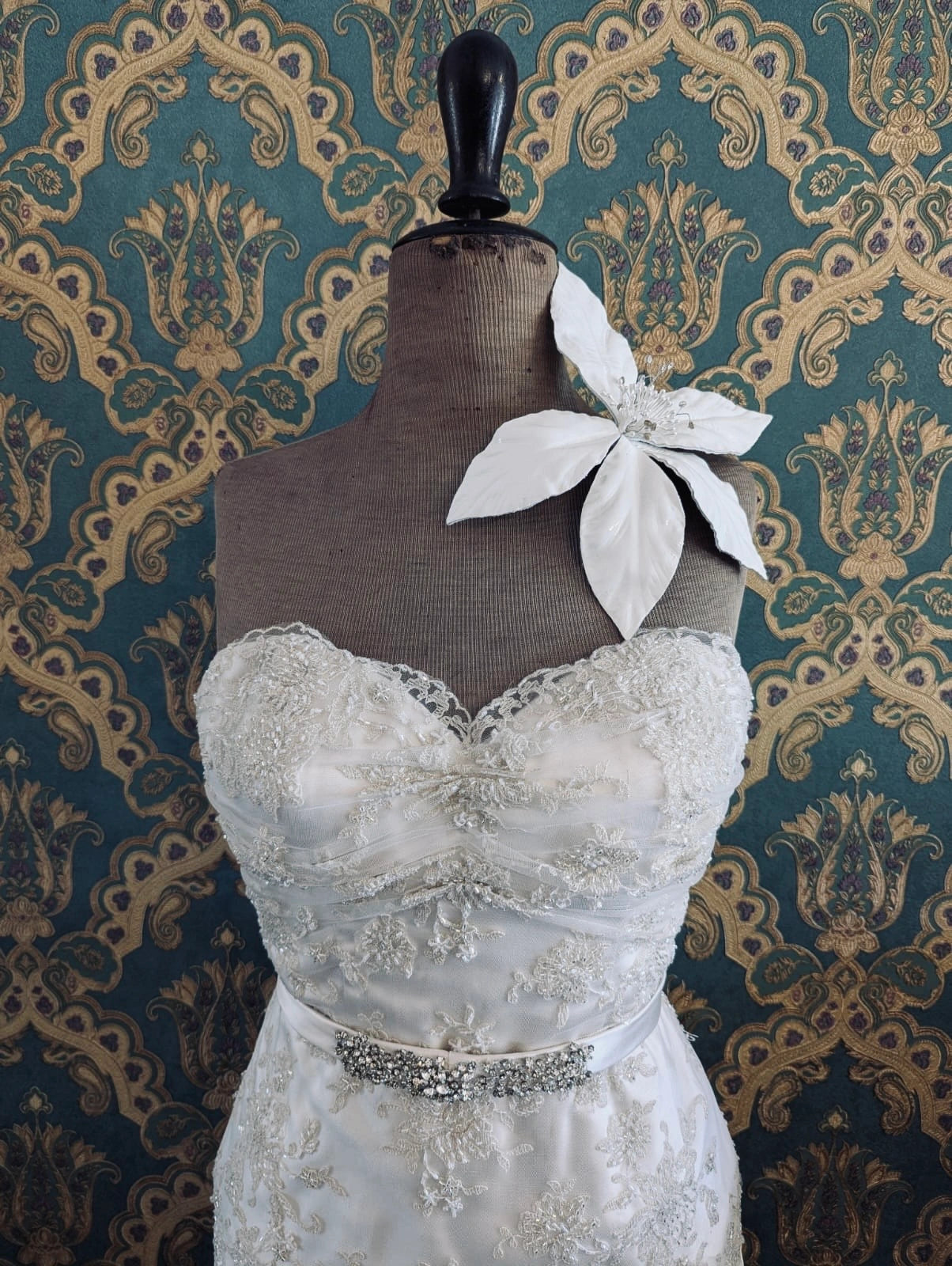 Broochella Giglio lily bridal flower brooch. Handmade in patent leather with a crystal spray from the centre. Pictured on a mannequin that is wearing a white, beaded strapless wedding dress. The background is green and gold damask wallpaper. 