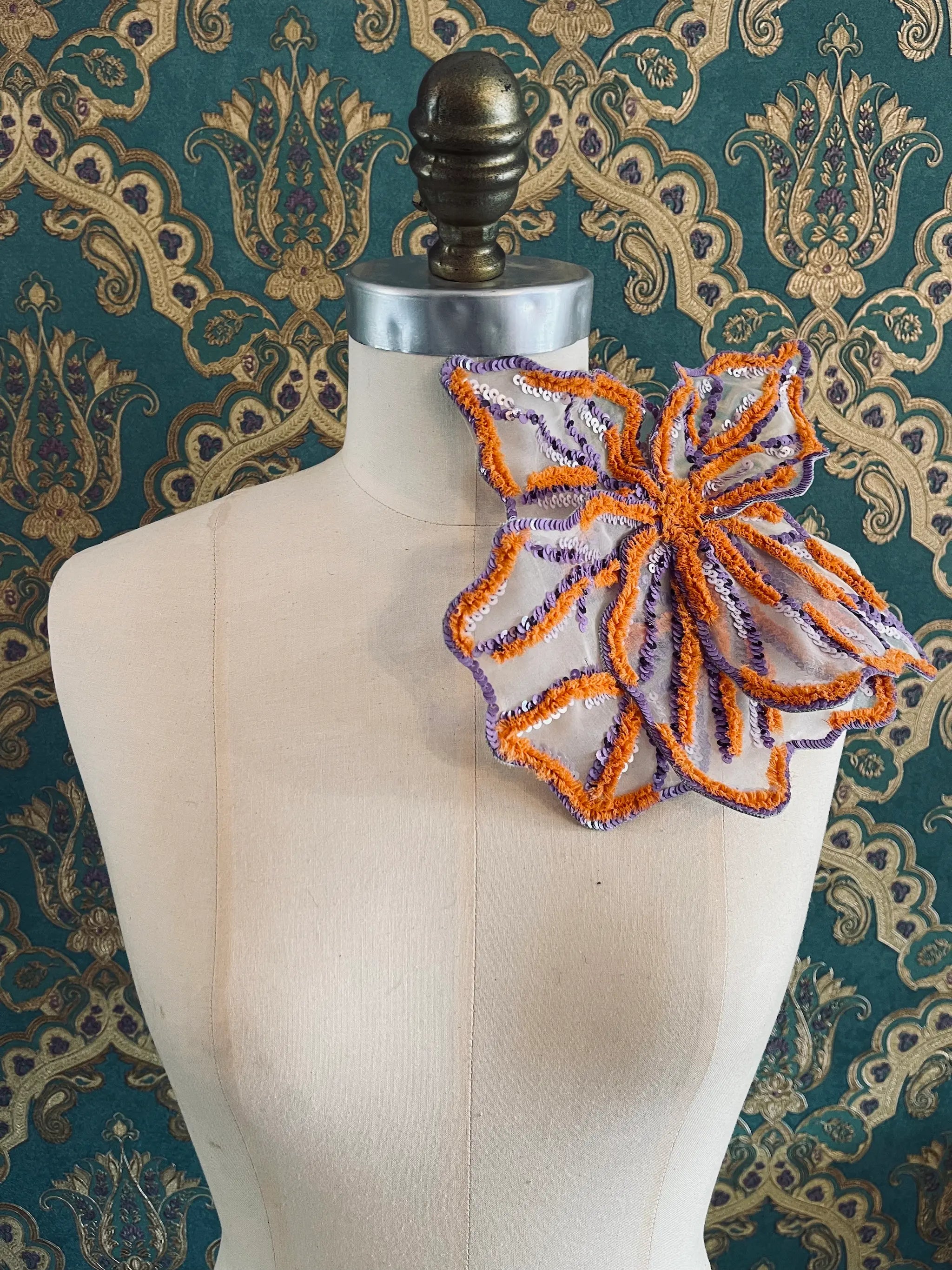 Broochella Giglio di tigre large handmade flower brooch in orange and purple. Pictured on a mannequin. The background is green and gold damask wallpaper