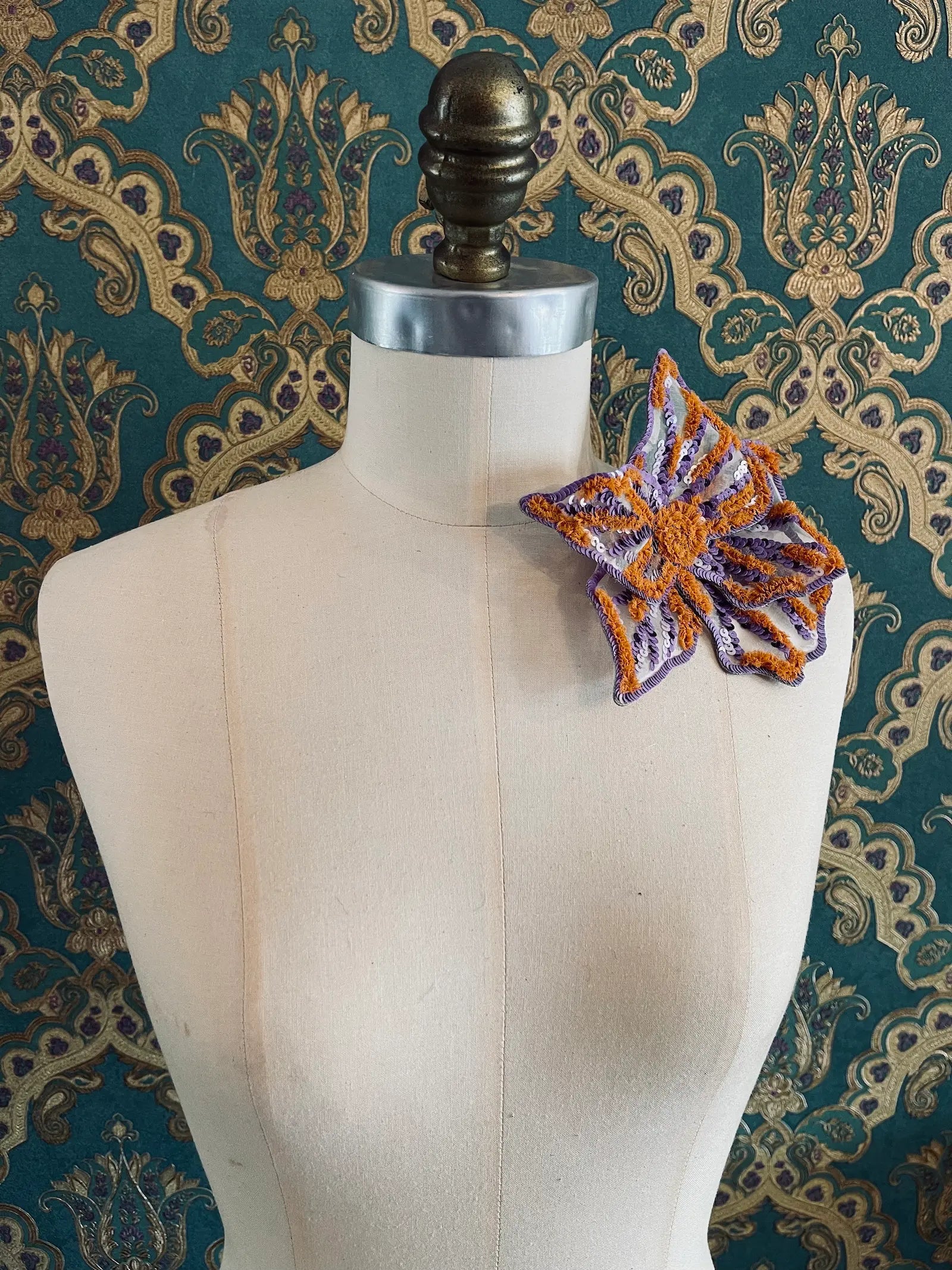Broochella giglio di tigre small hand beaded, sequinned, flower brooch in purple and orange. It is pinned to a mannequin. The background is green and gold damask wallpaper