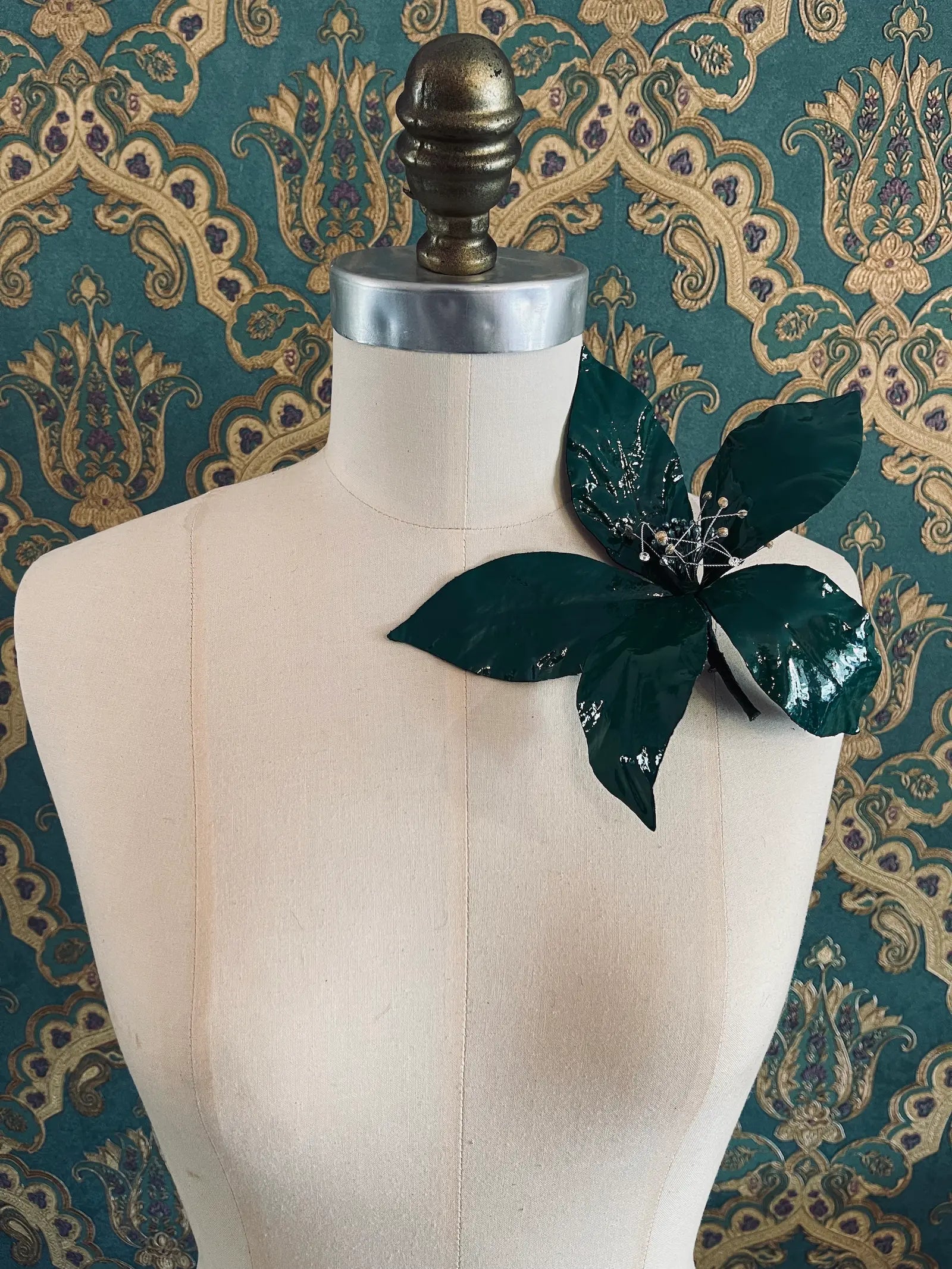 Broochella Giglio green patent leather large lily flower brooch. Handmade in New York. It is presented on a mannequin. The background is green and gold damask wallpaper.