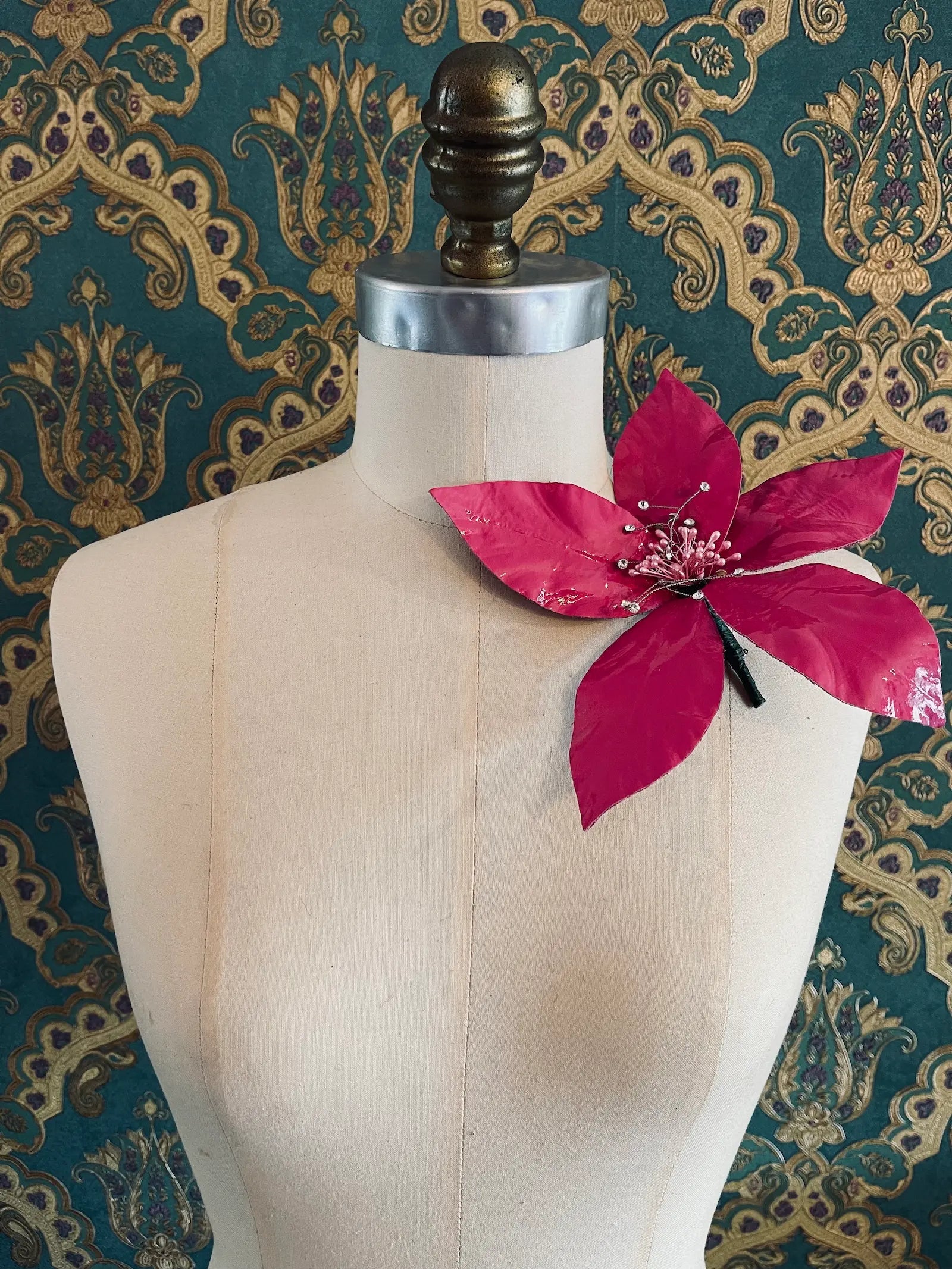 Broochella Giglio pink patent leather large lily flower brooch. Handmade in New York. It is presented on a mannequin. The background is green and gold damask wallpaper.