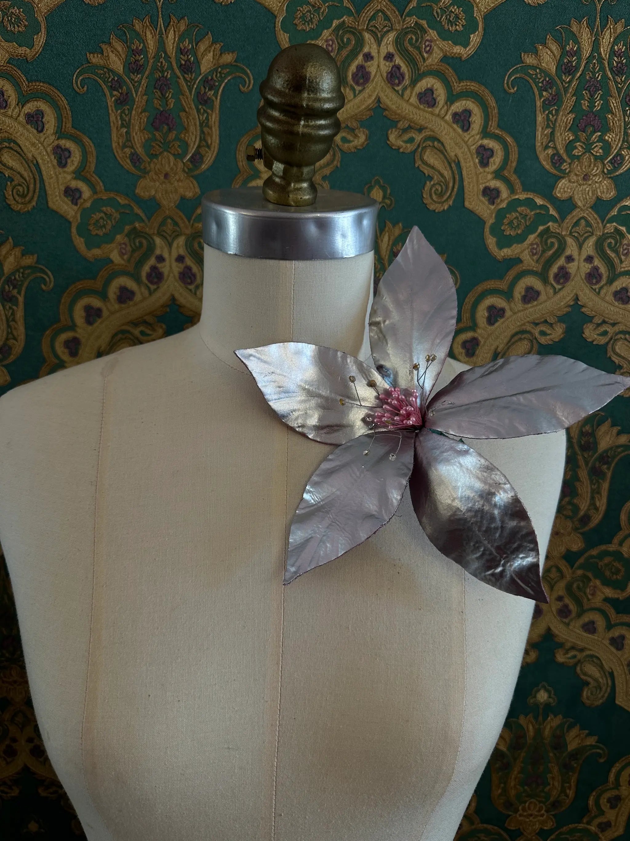 Broochella giglio handmade large patent leather flower in silver. Styled on a mannequin. The background is green and gold damask wallpaper