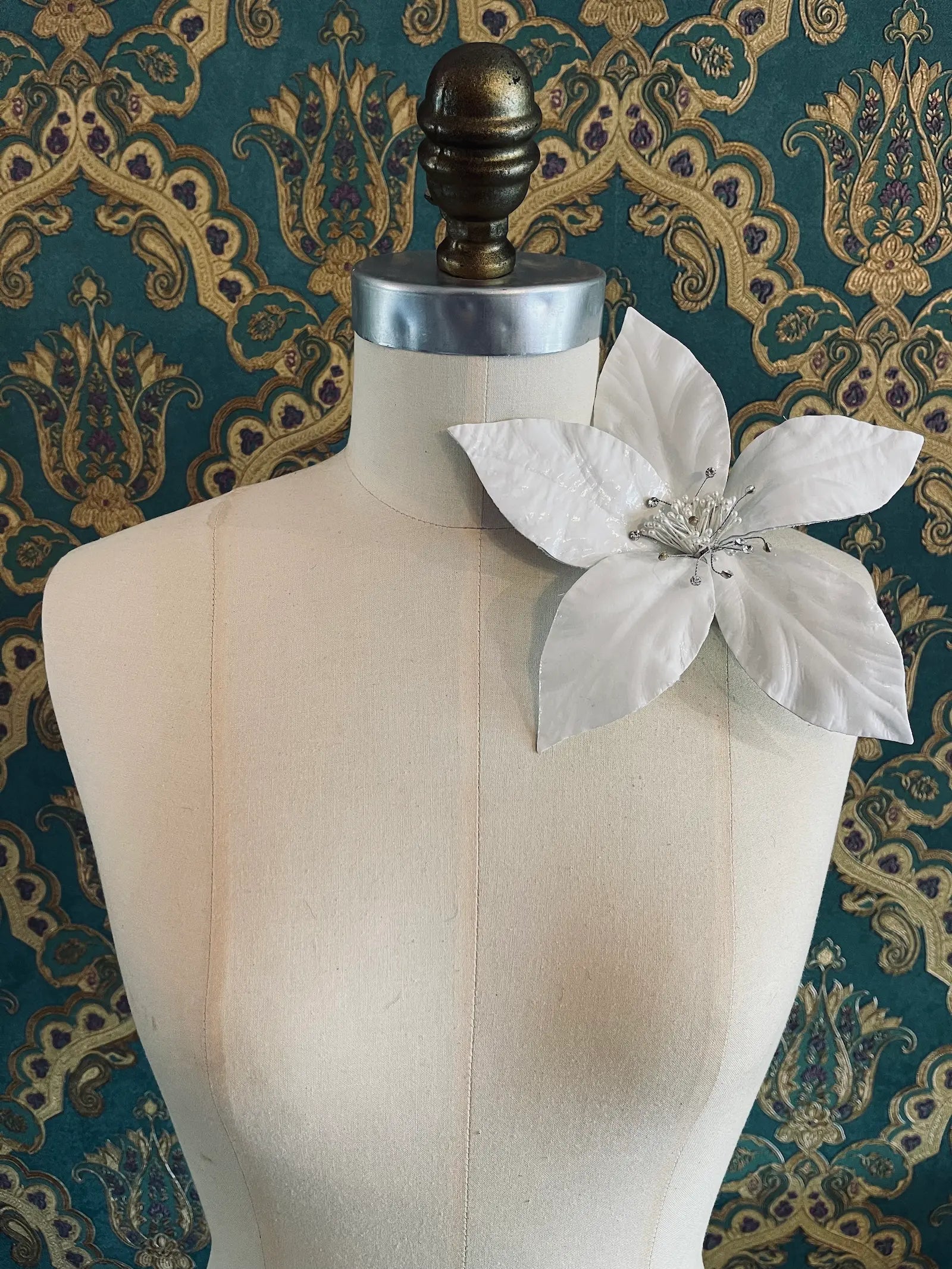 Broochella Giglio white patent leather large lily flower brooch. Handmade in New York. It is presented on a mannequin. The background is green and gold damask wallpaper.