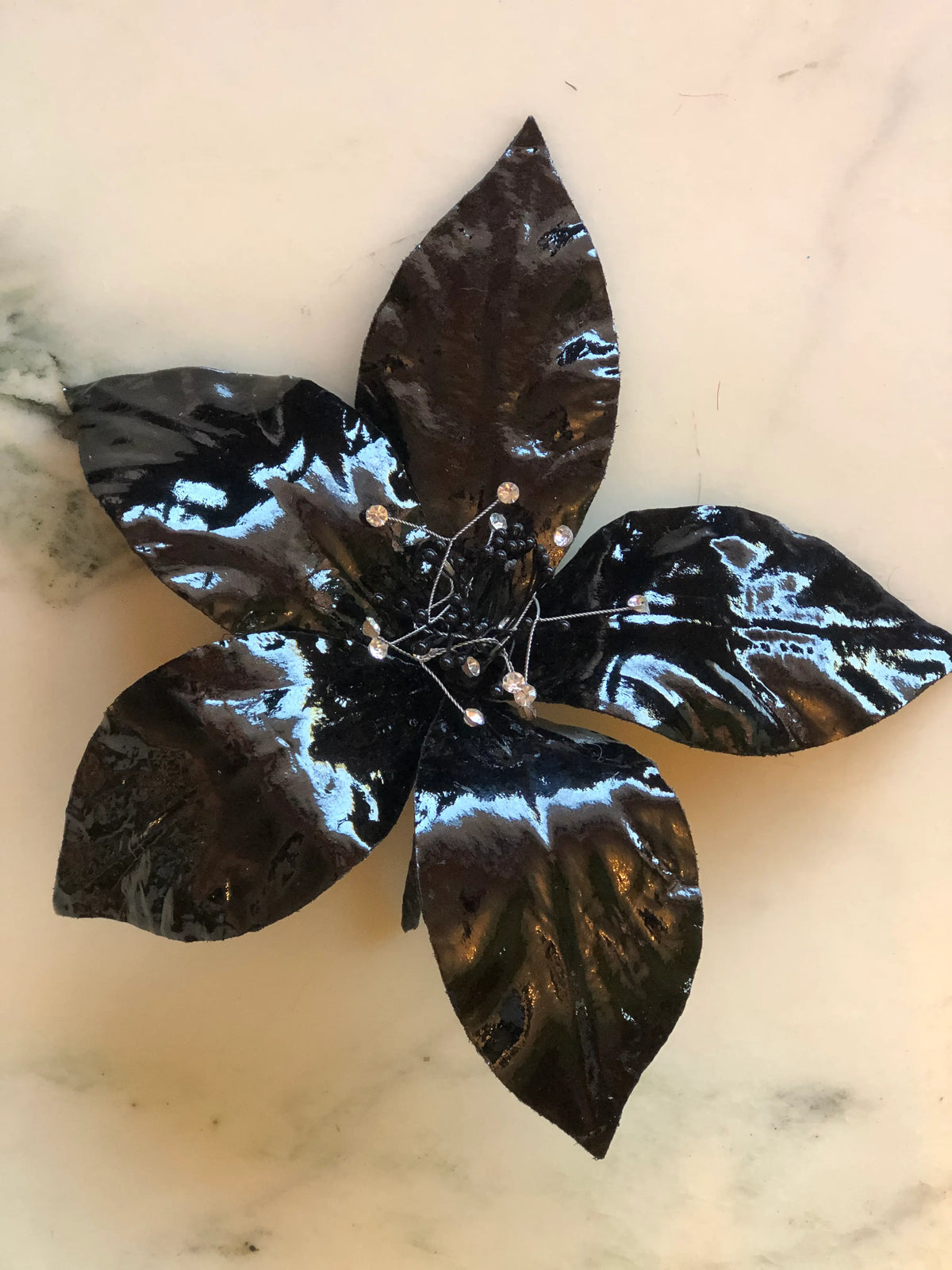 Broochella Men Giglio Flower Brooch Pin in glossy black, featuring sculptural leather lily petals with a textured black centre and delicate crystal-tipped stamens on a cream marble background.