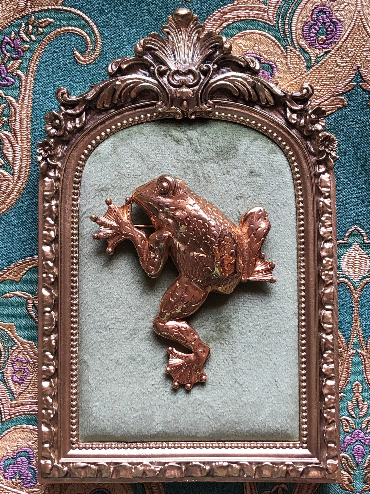 Broochella Gilded Frog vintage gold-tone statement brooch with a sculptural body, expressive eyes and detailed webbed feet, measuring approximately 8.5 × 7 cm. Photographed on a velvet tray with an ornate gold frame. The background is green and gold damask wallpaper