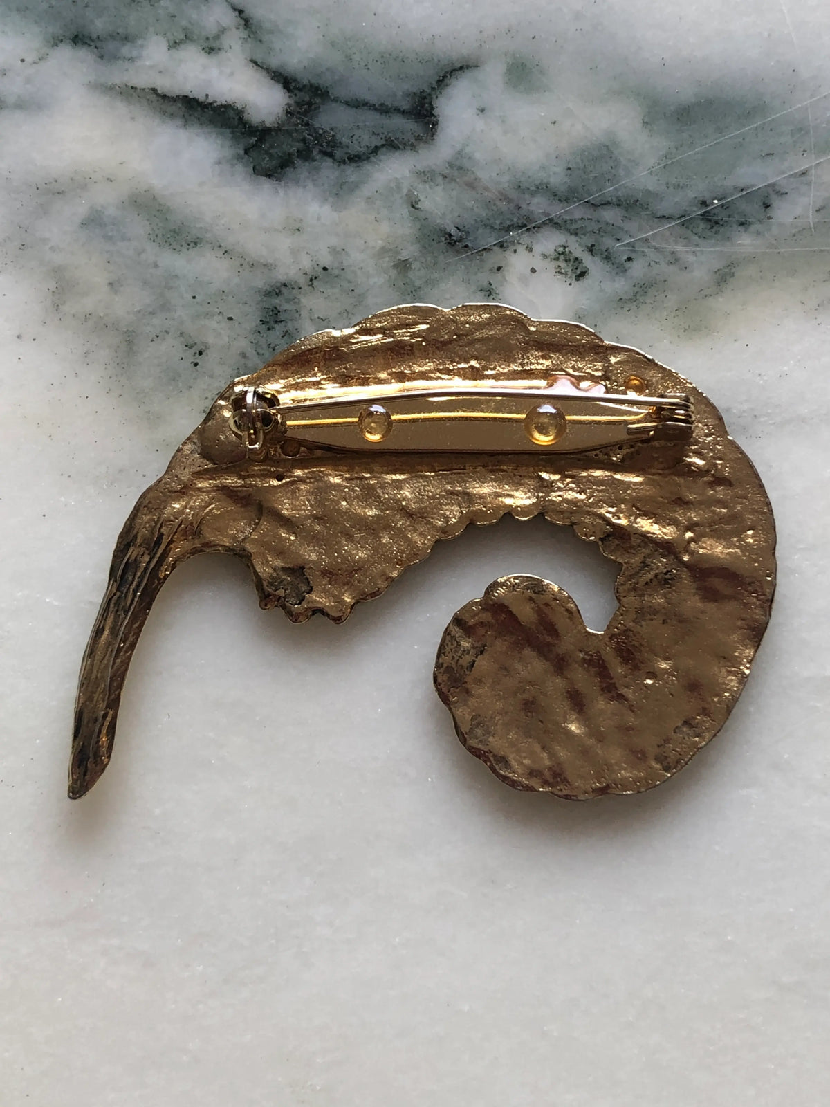 Broochella Gilded Plume vintage feather brooch with an antiqued gold-tone finish, curled sculptural shape and deeply engraved detailing, measuring approximately 5 × 7 cm. Photographed on white and grey marble.