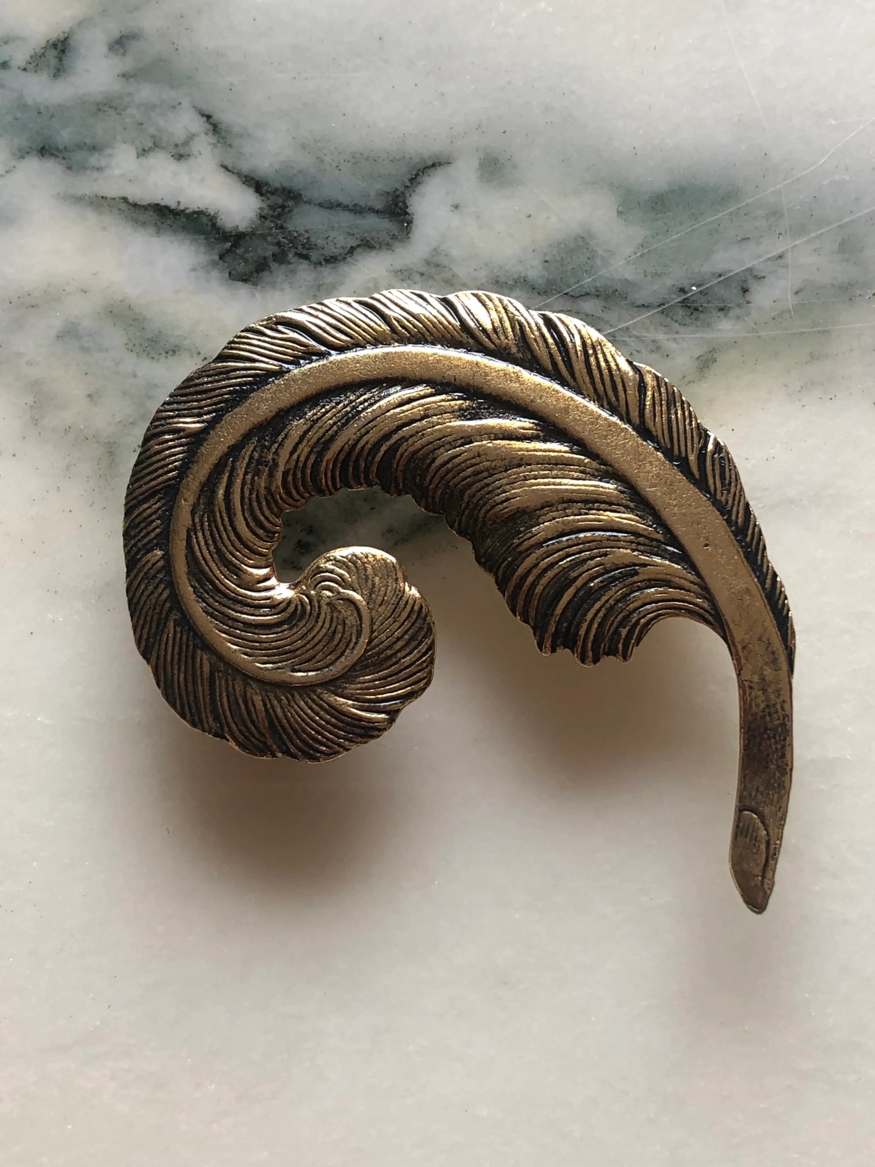 Broochella Gilded Plume vintage feather brooch with an antiqued gold-tone finish, curled sculptural shape and deeply engraved detailing, measuring approximately 5 × 7 cm. Photographed on white and grey marble.