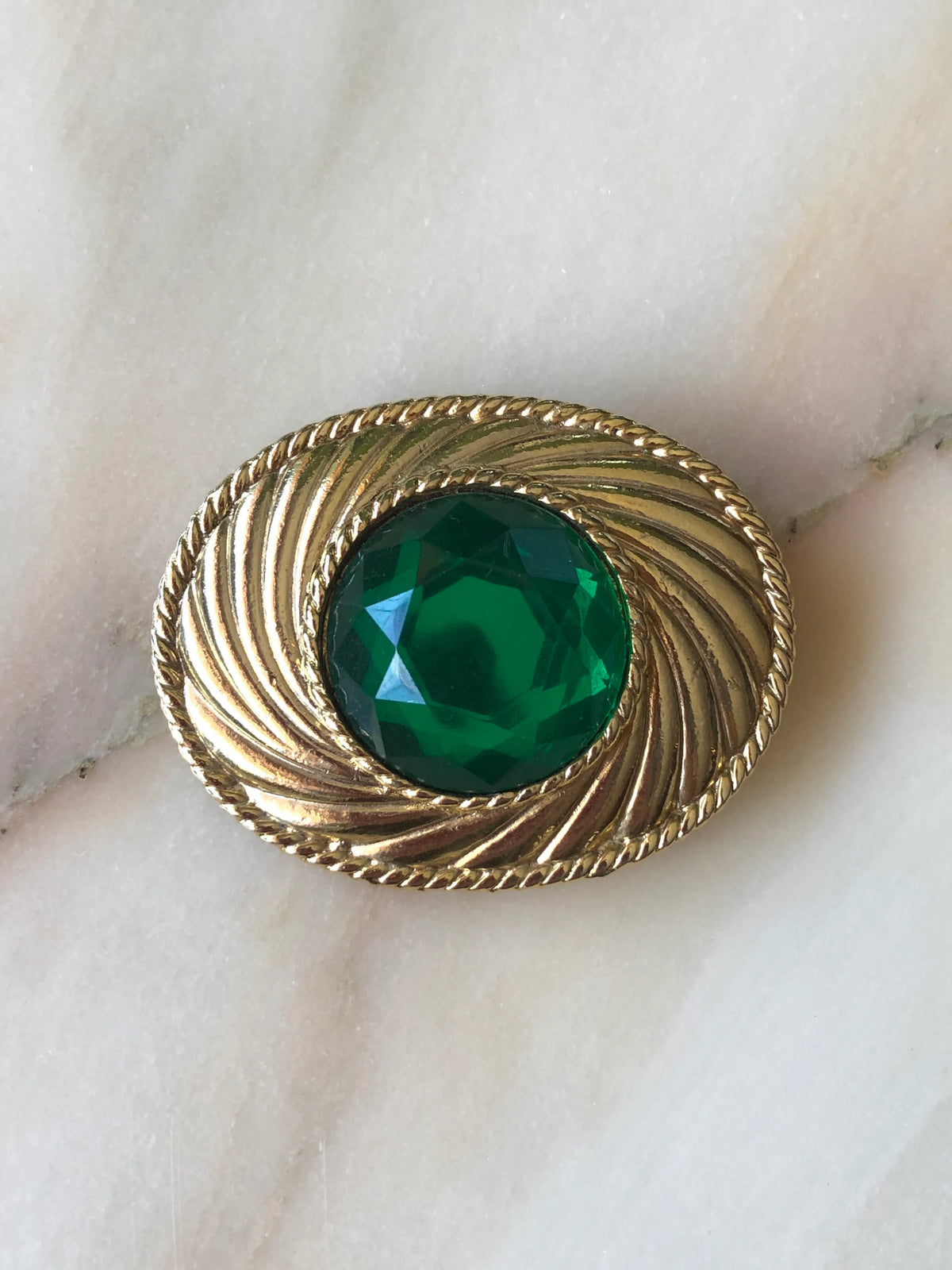 Broochella Giulia vintage Ungaro oval brooch featuring a large faceted emerald green glass centrepiece, surrounded by sculpted gold-tone swirls and a twisted rope border, photographed on a cream marble background with soft beige veining.