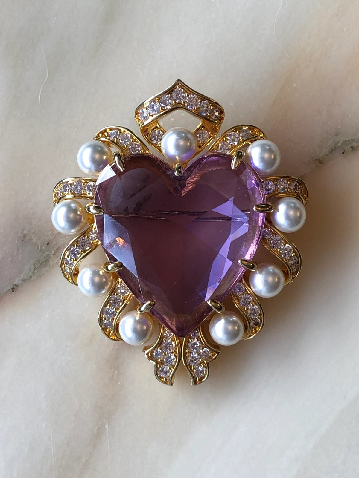 A Broochella Giulietta e Romeo heart-shaped brooch featuring a faceted purple heart gemstone surrounded by white pearls and gold settings with small clear crystals, displayed on a light marble surface.