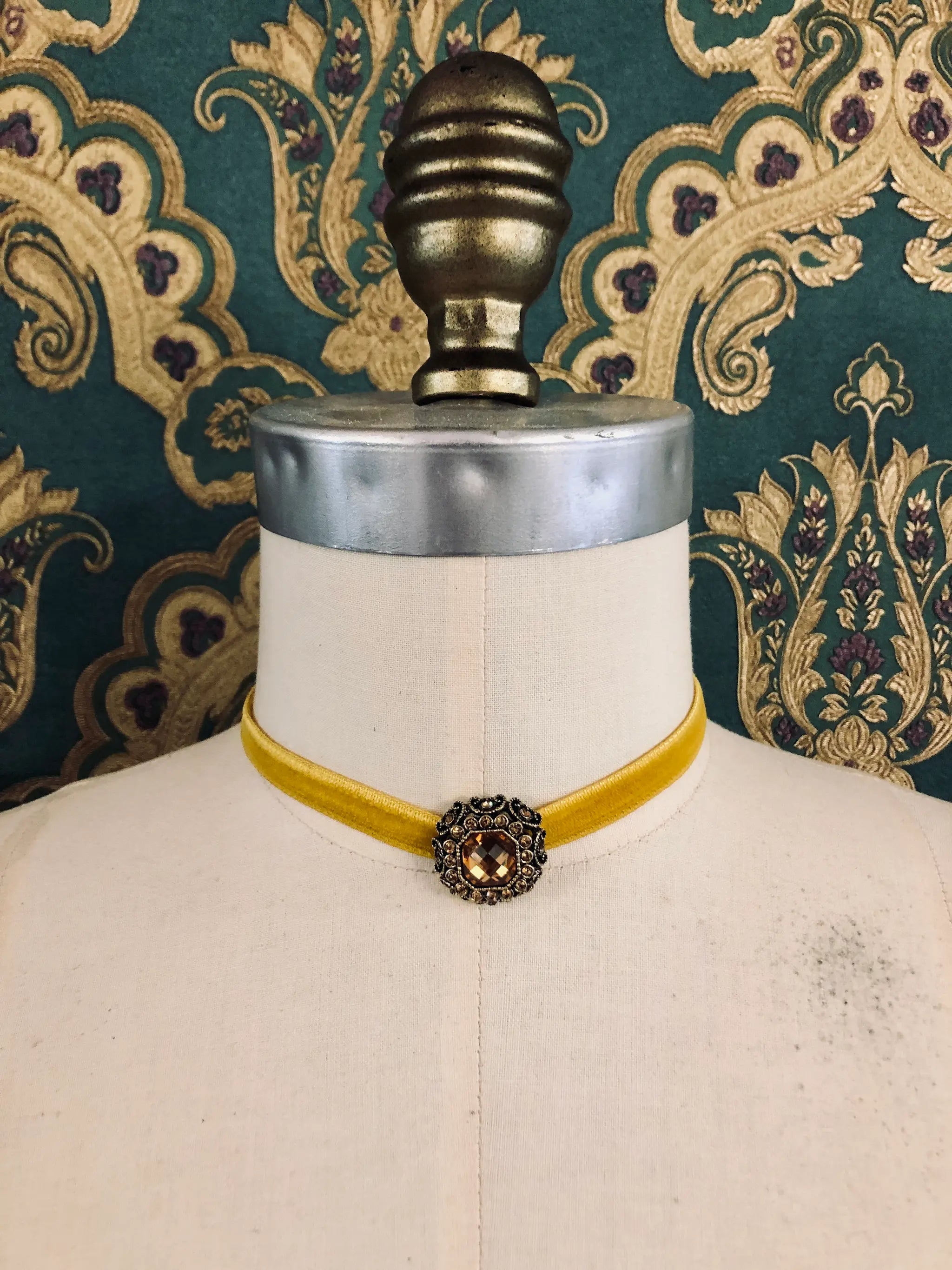 A Broochella Lady Whistledown mustard yellow velvet choker necklace with a decorative antique bronze filigree centrepiece, displayed on a cream-coloured dress form against a patterned teal and gold damask wallpaper background.