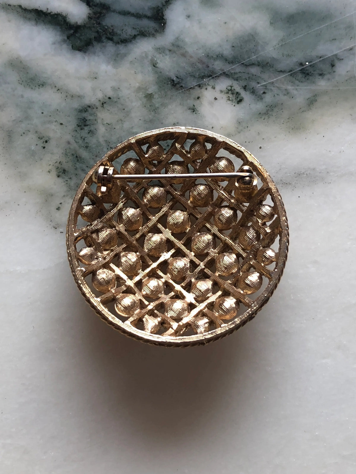 The back view of Broochella Golden Constellation vintage round brooch with raised gold-tone discs and an intricate twisted rope lattice, measuring approximately 4 cm across. Photographed on white and grey marble