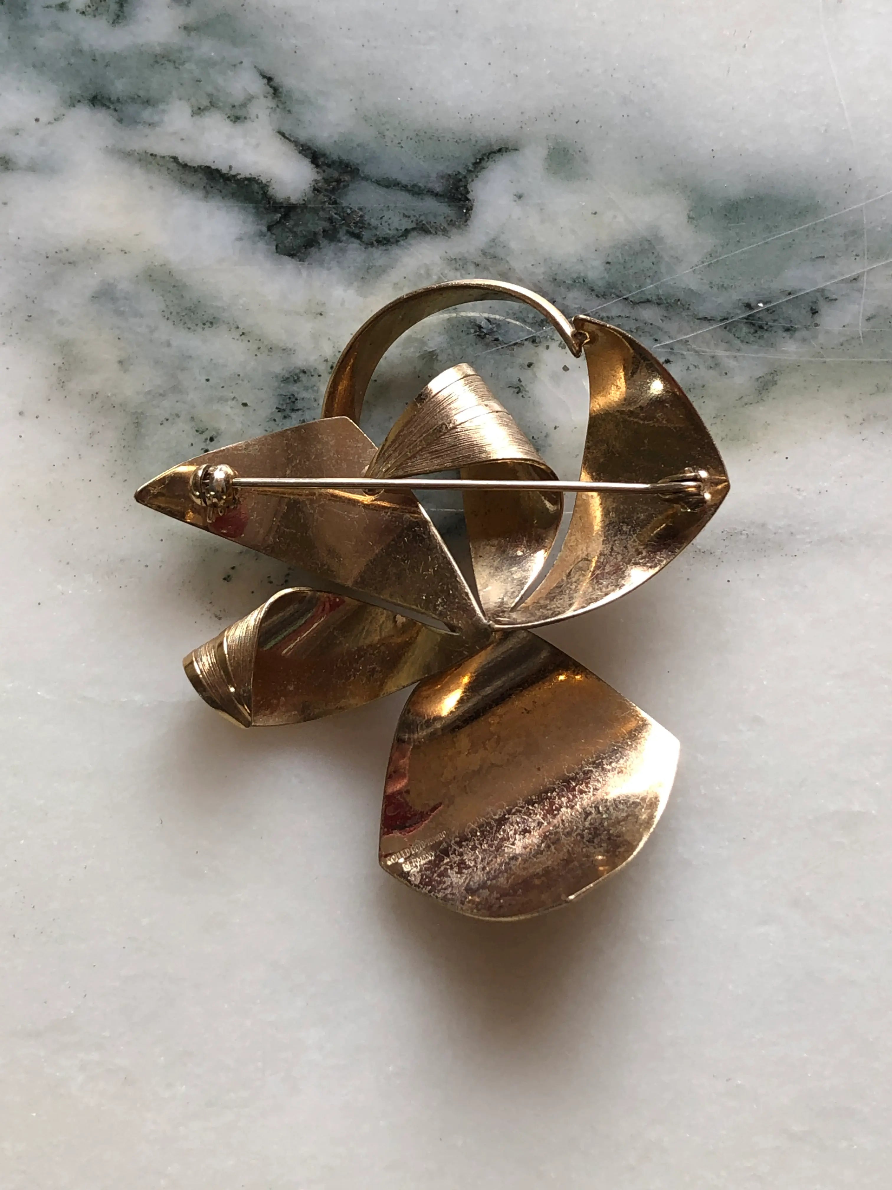 Broochella Golden Flourish vintage gold-tone brooch with an abstract bow, sweeping ribbon shapes and fine textured detailing, measuring approximately 6 × 5 cm. Photographed on white and grey marble.