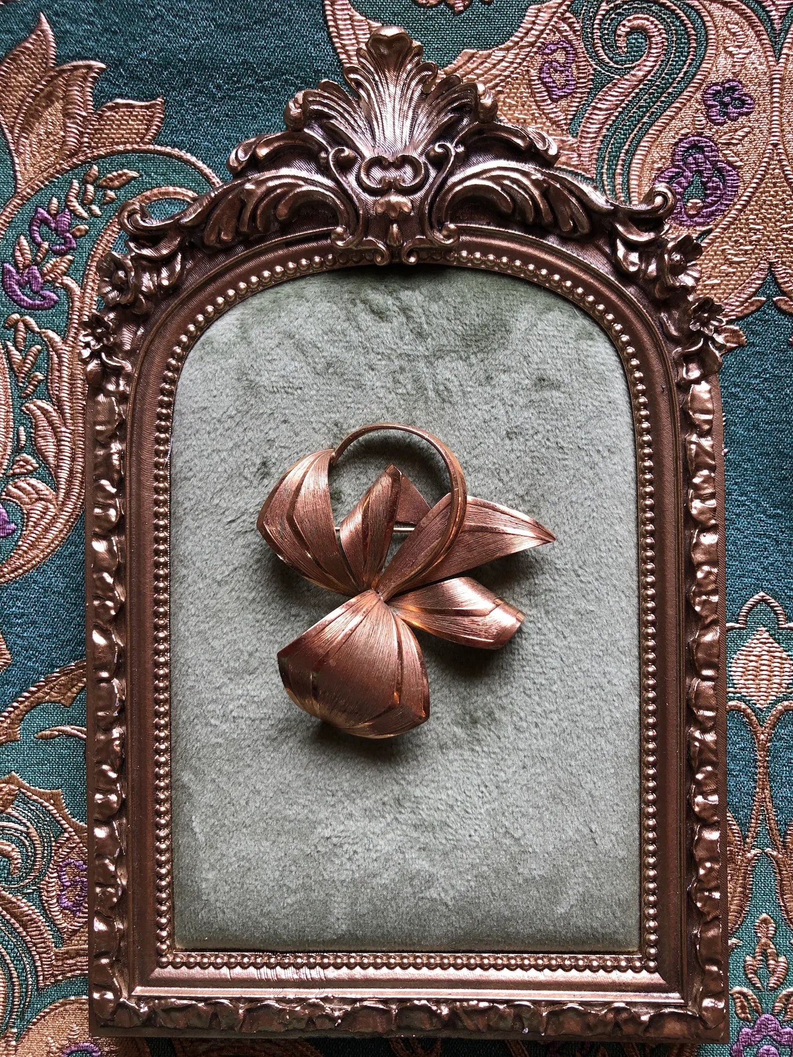 Broochella Golden Flourish vintage gold-tone brooch with an abstract bow, sweeping ribbon shapes and fine textured detailing, measuring approximately 6 × 5 cm. Photographed on a green velvet tray with an ornate, gold frame. The background is green and gold damask wallpaper