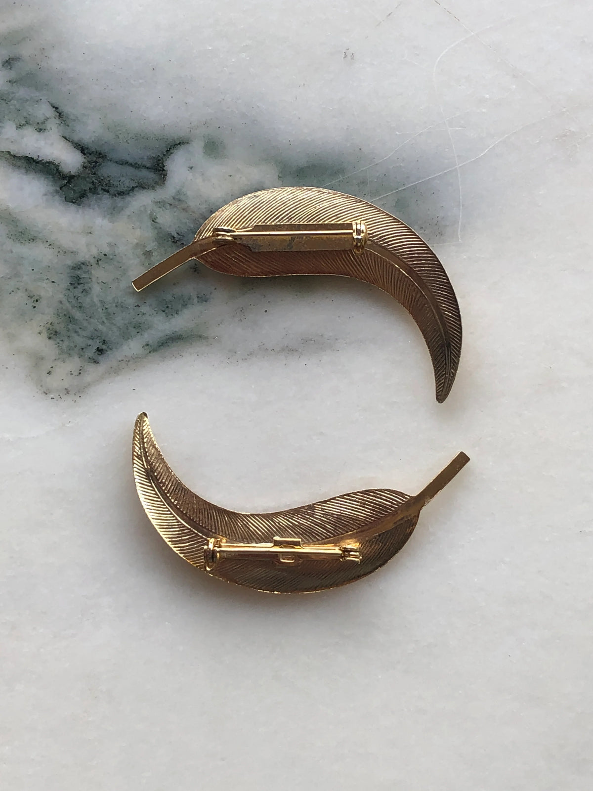 Broochella Golden Gum Leaf Vintage Brooch Pair back view showing the textured gold-tone leaves and traditional pin fastenings, photographed on a white marble background with soft grey-green veining.
