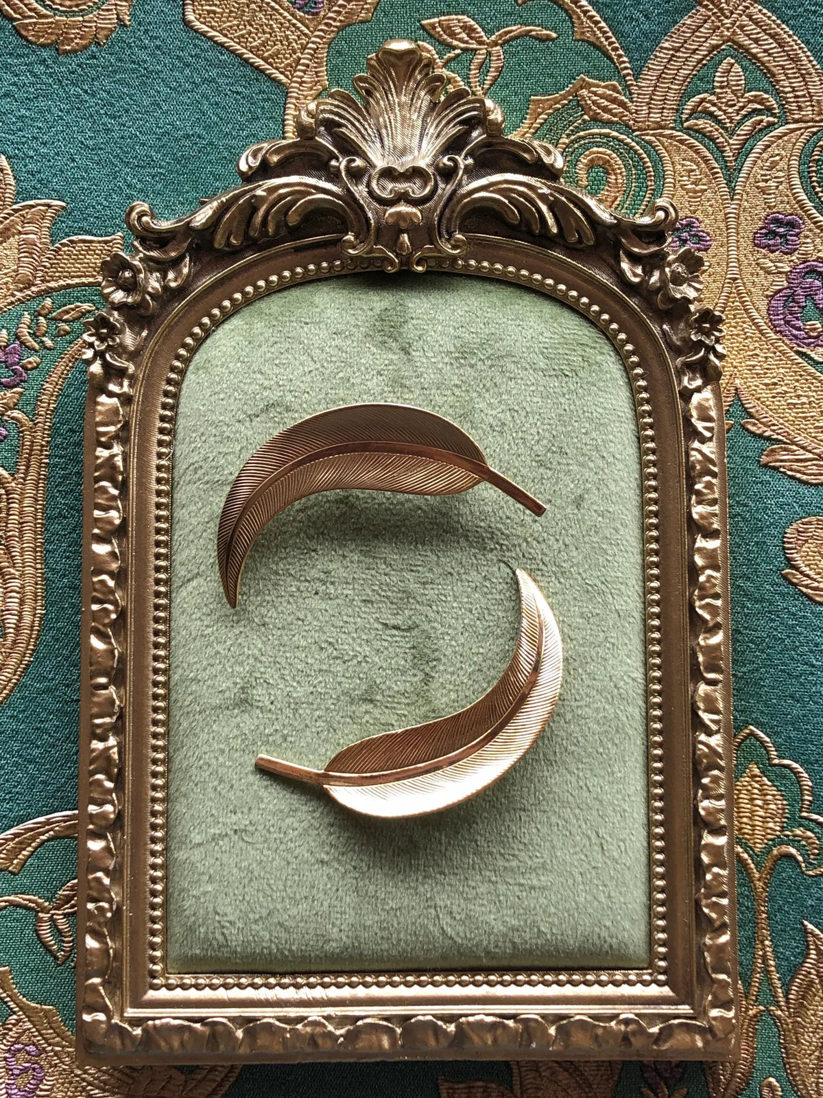 Broochella Golden Gum Leaf Vintage Brooch Pair featuring two curved gold-tone gum leaf brooches with finely textured leaf detailing, displayed on sage green velvet in an ornate antique gold frame against green and gold patterned fabric.