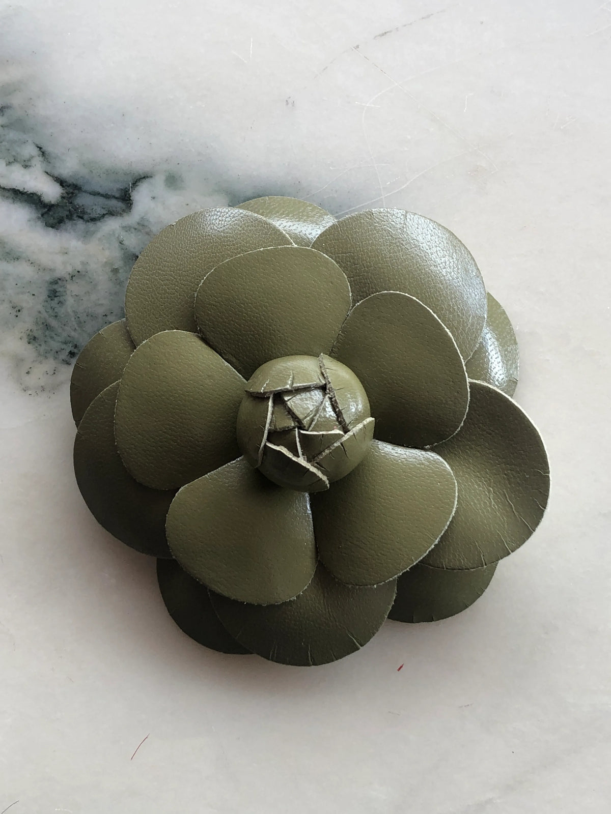 Broochella Camelia Leather Flower Brooch in olive green, featuring handcrafted layered kid-skin leather petals and a sculptural flower centre on a cream marble background.