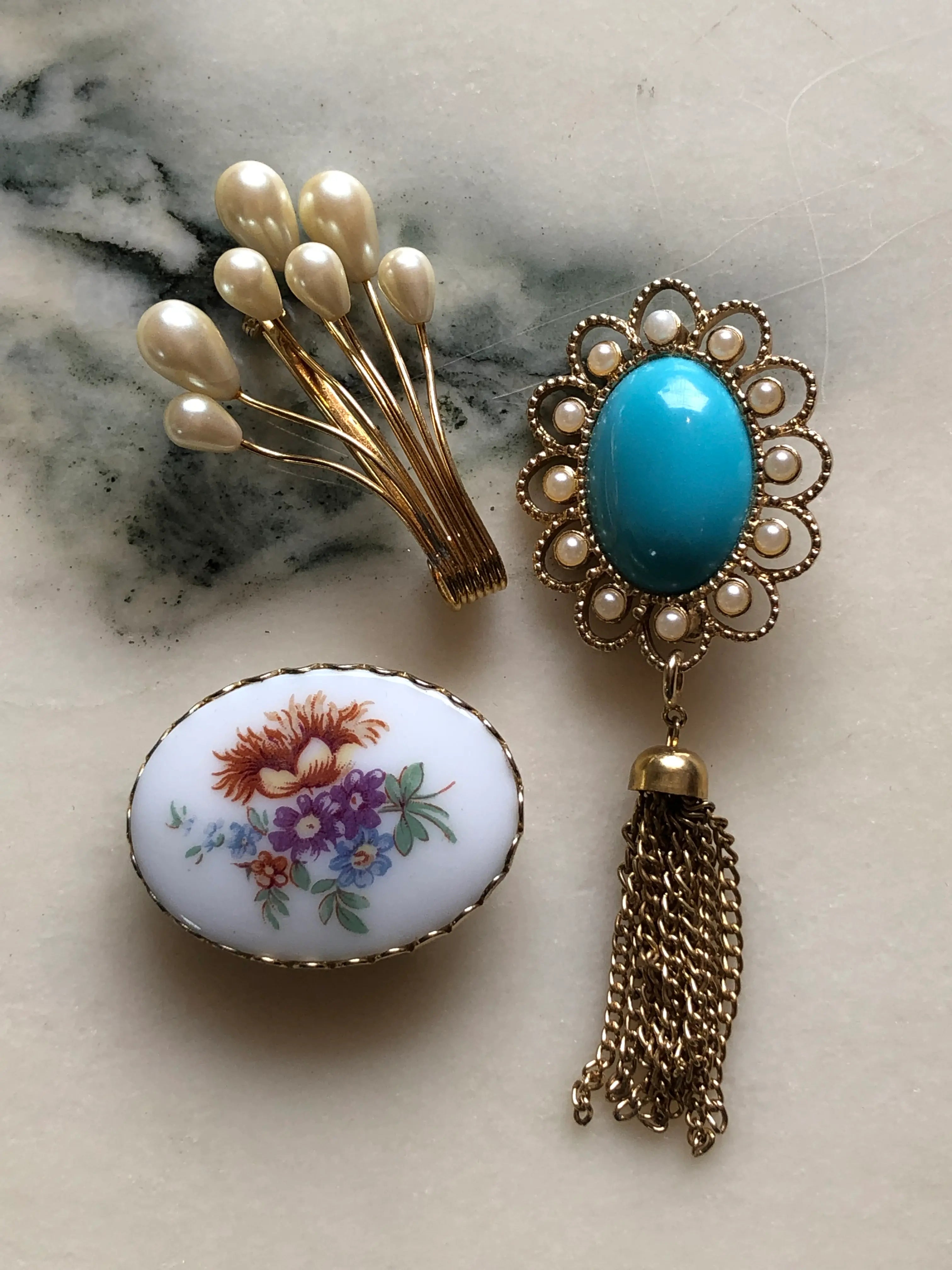 Broochella The Heirloom Trio vintage brooch set featuring a gold-tone faux-pearl spray, an oval floral brooch and an ornate turquoise-blue cabochon brooch with faux pearls and a chain tassel. Photographed on white and grey marble.