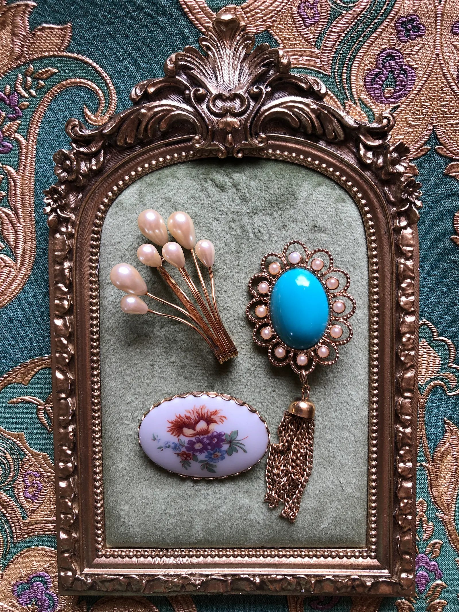Broochella The Heirloom Trio vintage brooch set featuring a gold-tone faux-pearl spray, an oval floral brooch and an ornate turquoise-blue cabochon brooch with faux pearls and a chain tassel. Photographed on a green velvet tray with an ornate gold frame. The background is green and gold damask wallpaper