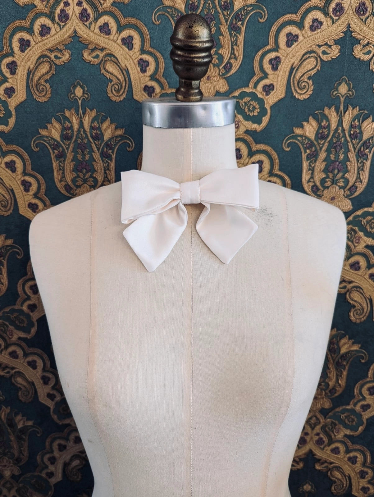 Broochella hollywood handmade satin bow in ivory. The brooch is pinned to a mannequin at the neck. The background is green and gold damask wallpaper.