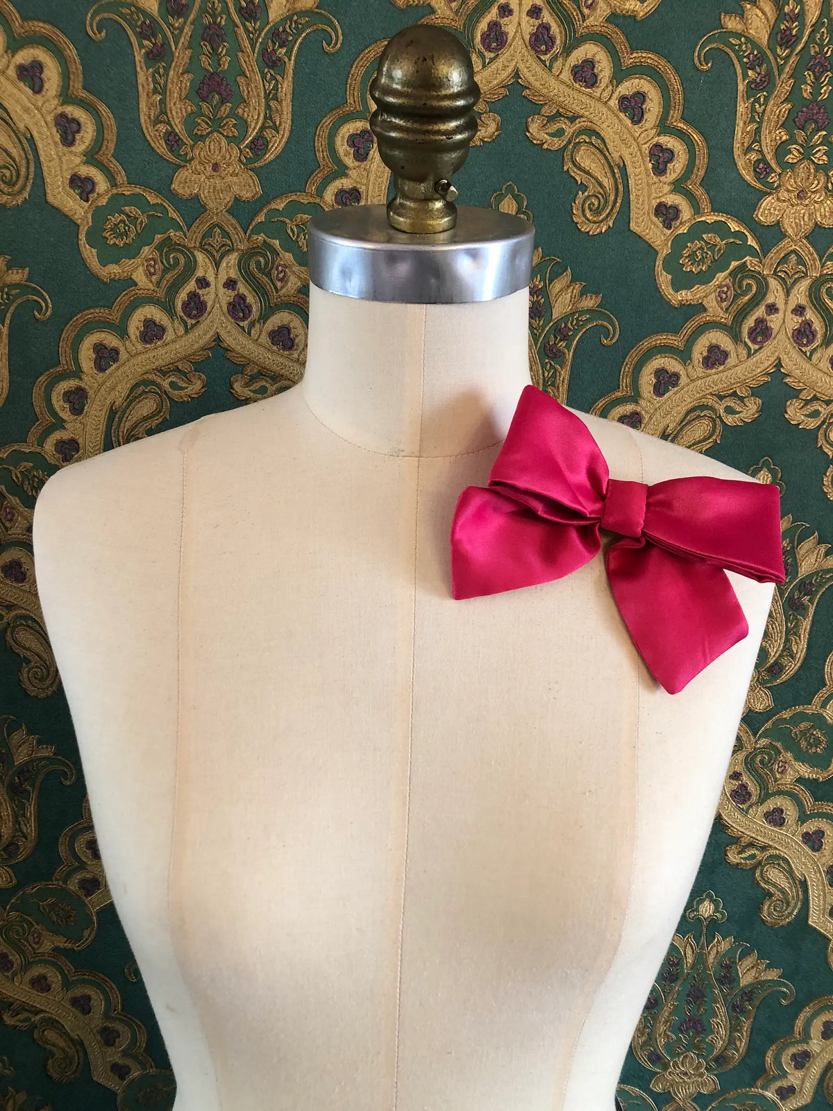 Broochella The Hollywood Satin Bow Brooch in vibrant pink styled on a mannequin against an ornate green and gold patterned background.