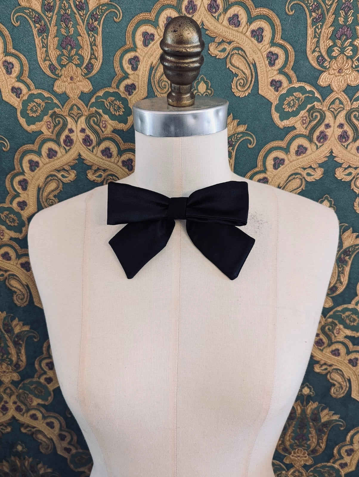 Broochella hollywood handmade satin bow in black. The brooch is pinned to a mannequin at the neck. The background is green and gold damask wallpaper.