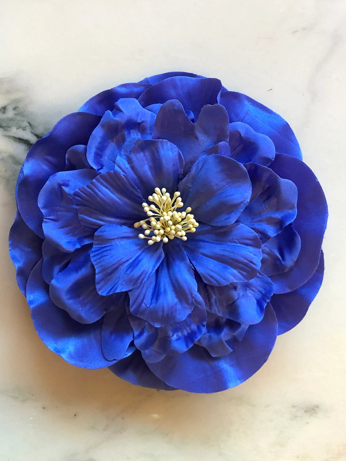 Broochella Ibisco Men's Flower Brooch in vivid cobalt blue, featuring full layered fabric petals with sculptural texture and a contrasting cream stamen centre on a cream marble background.