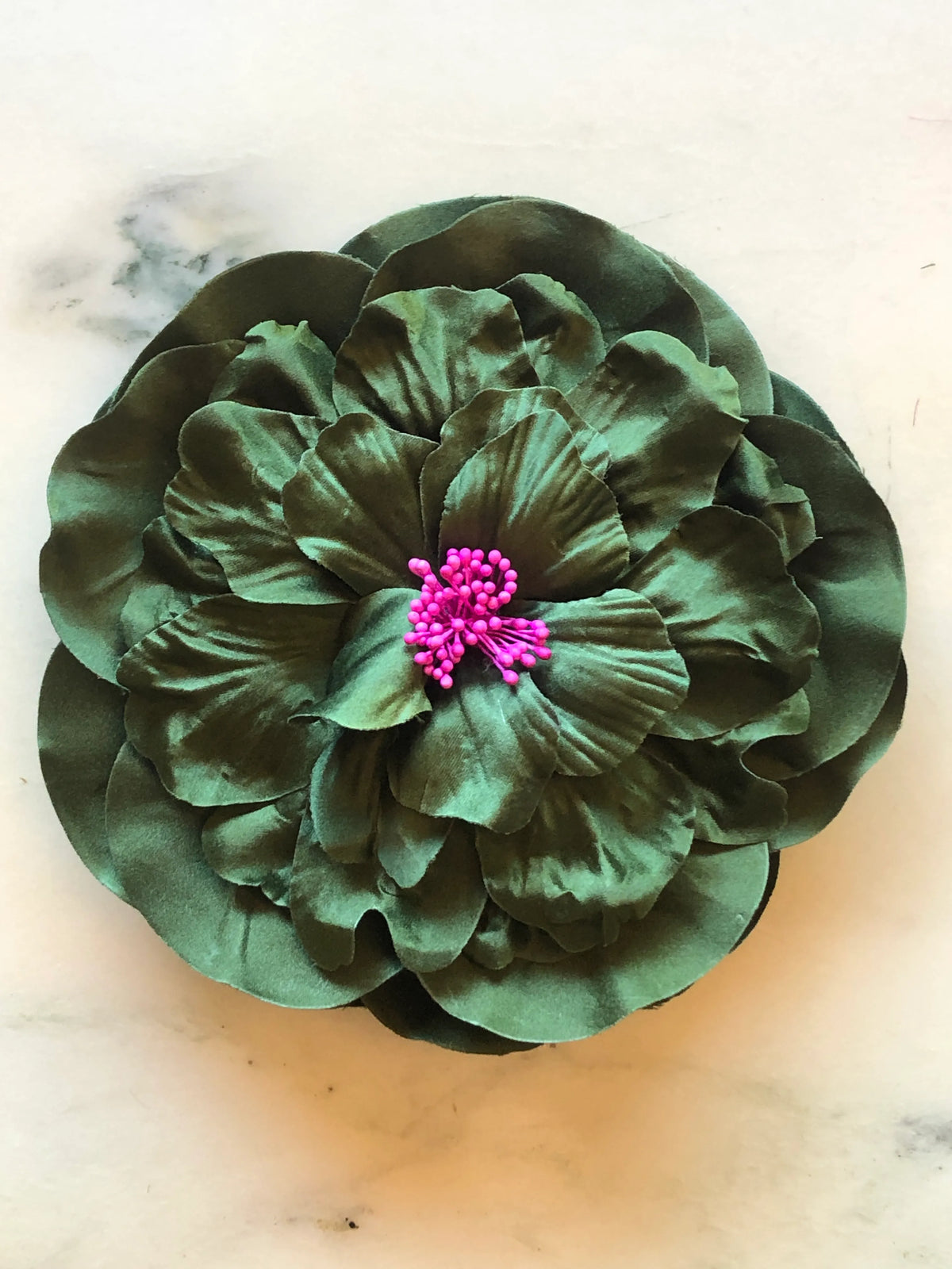 Broochella Men's Ibisco Flower Brooch in rich green, featuring full layered fabric petals with sculptural texture and a contrasting bright pink stamen centre on a cream marble background.