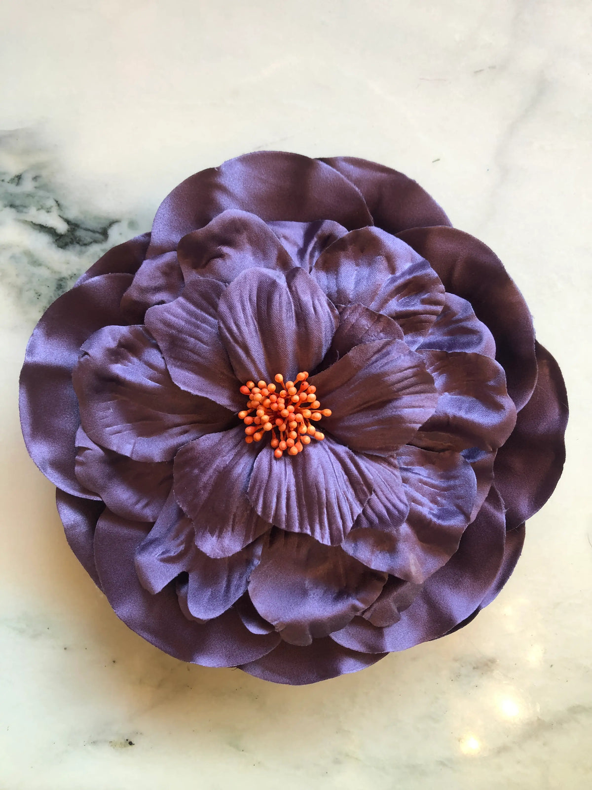Broochella Ibisco Flower Brooch in soft purple, featuring full layered fabric petals with sculptural texture and a contrasting orange stamen centre on a cream marble background.