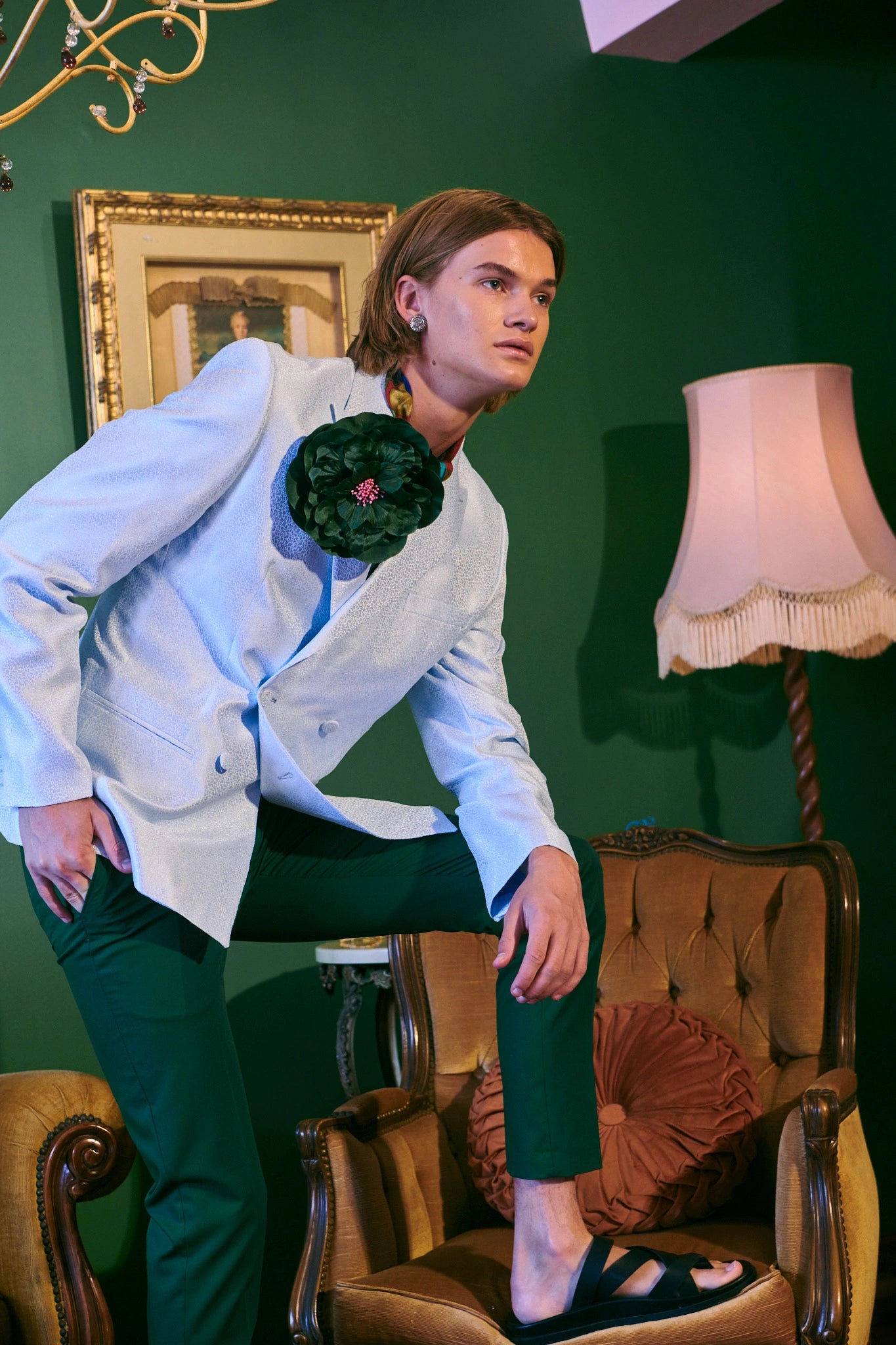 Man in a white coat and green pants posing in an ornate room with a lamp and framed picture on the wall. They are wearing the green Broochella ibisco flower brooch