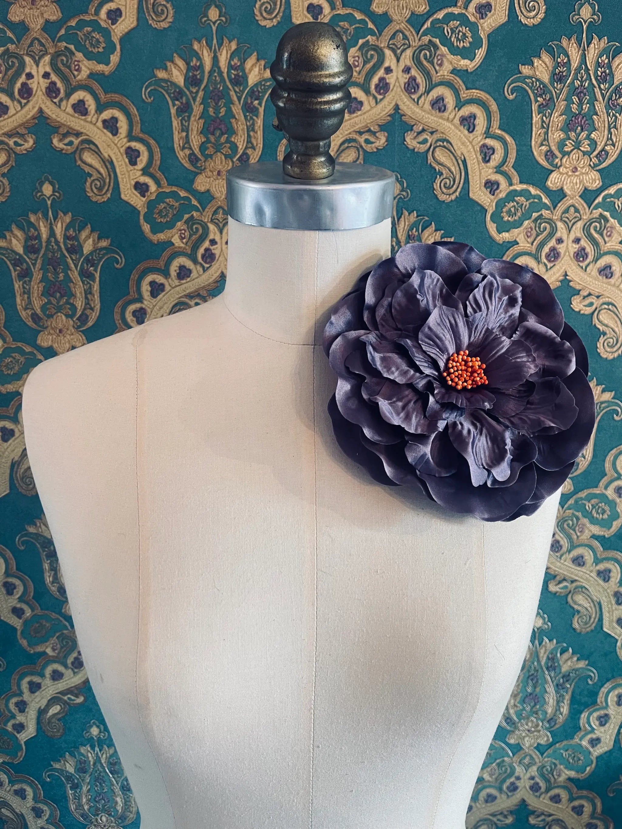 Broochella Ibisco large, purple, handmade flower brooch. Styled on a mannequin. The background is green and gold damask wallpaper