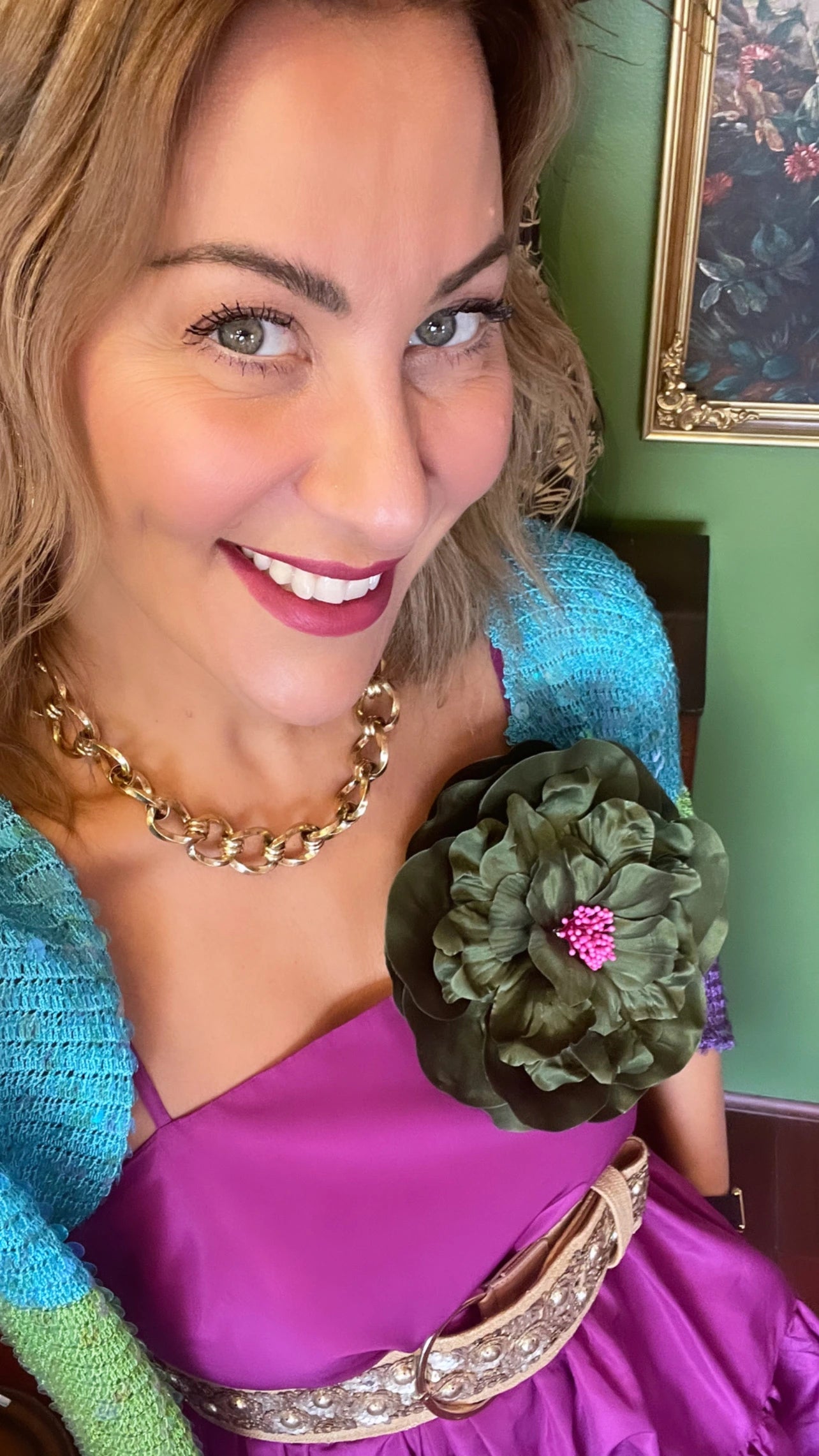 A smiling woman wearing a bright purple dress with a turquoise cardigan, a chunky gold chain necklace, and a large Broochella ibisco olive green fabric flower brooch with a pink centre.