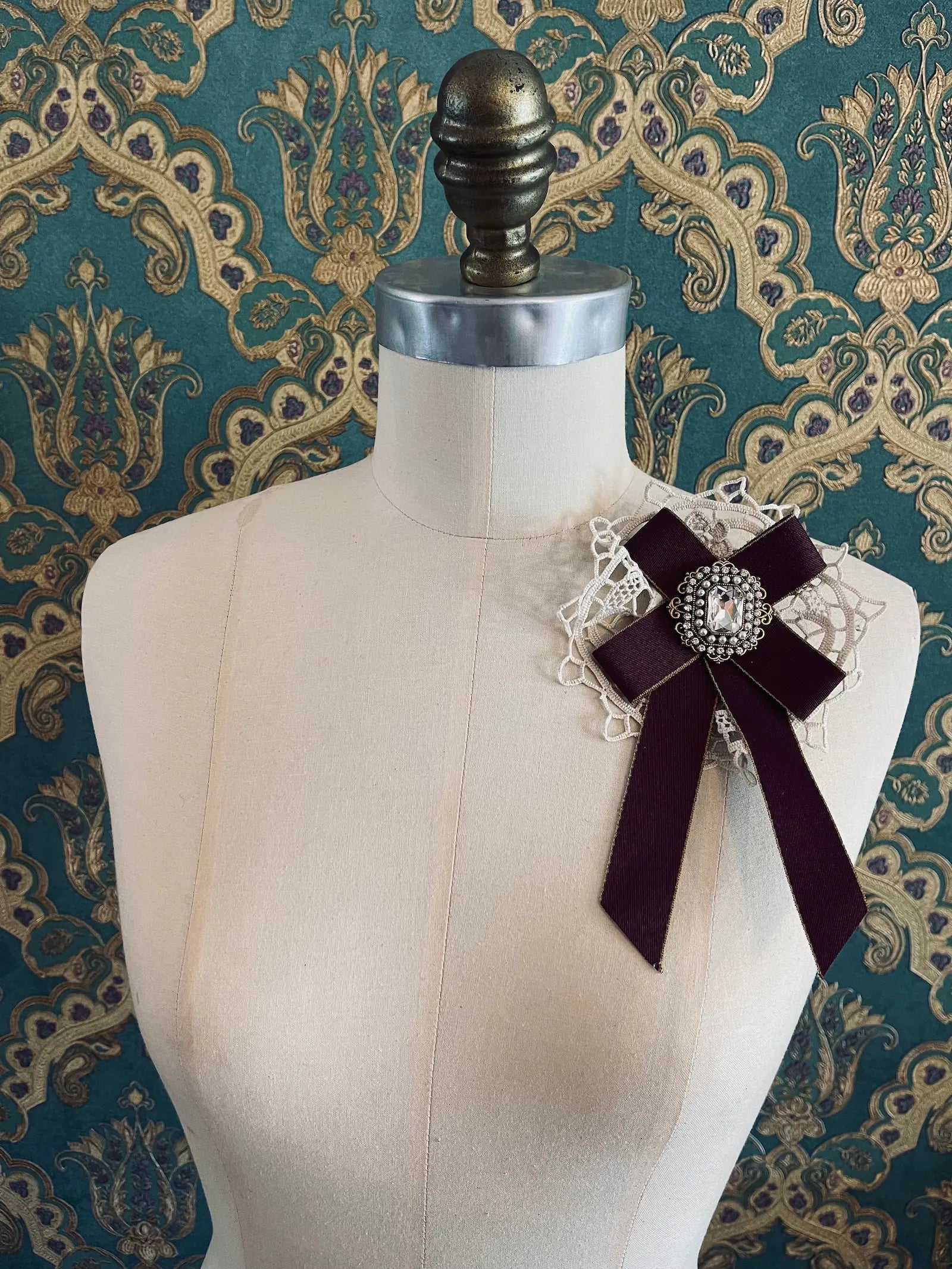 Broochella Il Conte burgundy handmade bow brooch with rhinestone centre embellishment. Pictured on a mannequin. The background is green and gold damask wallpaper