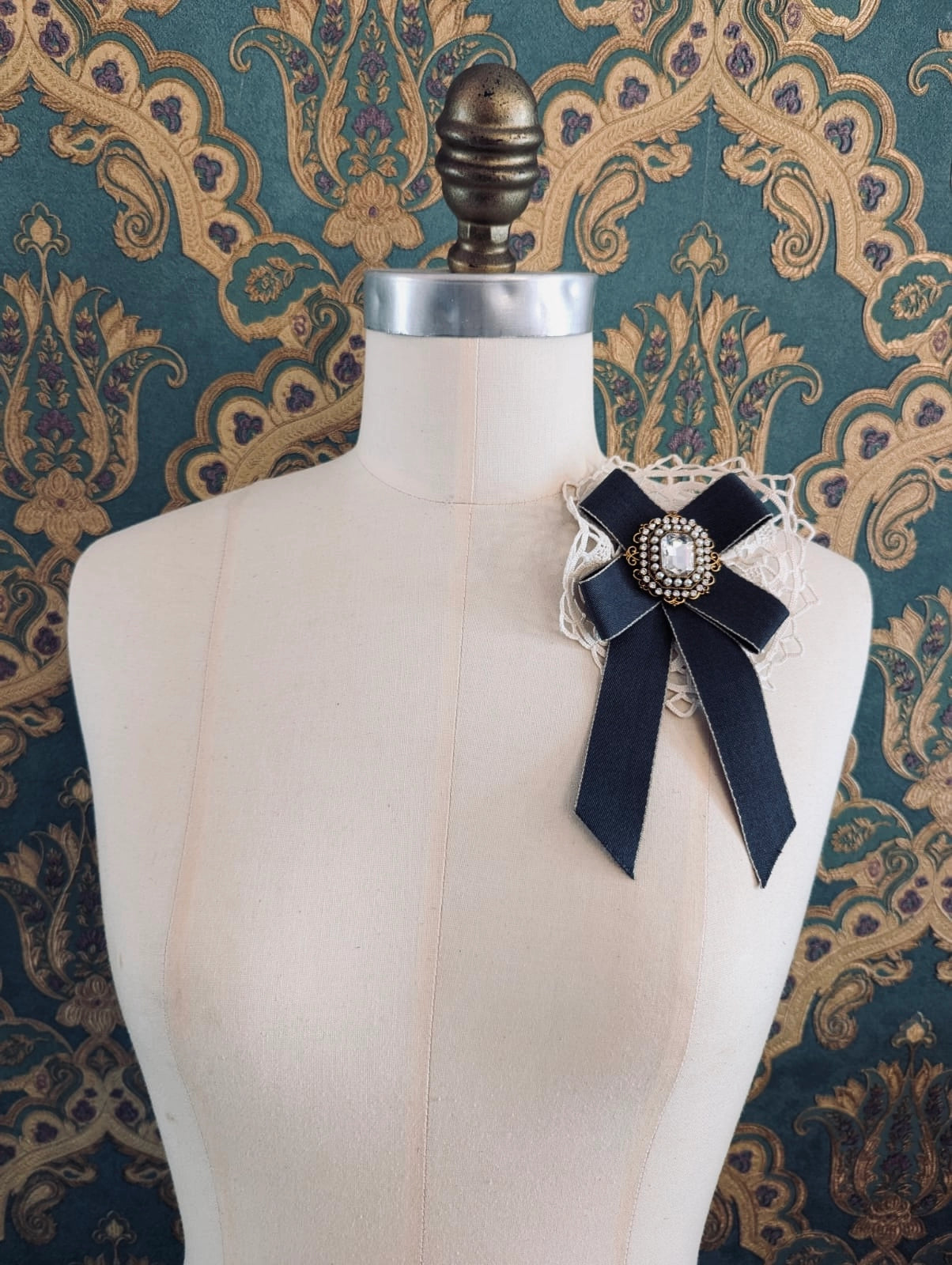 Broochella Il Conte grey handmade bow brooch with rhinestone centre embellishment. Pictured on a mannequin. The background is green and gold damask wallpaper