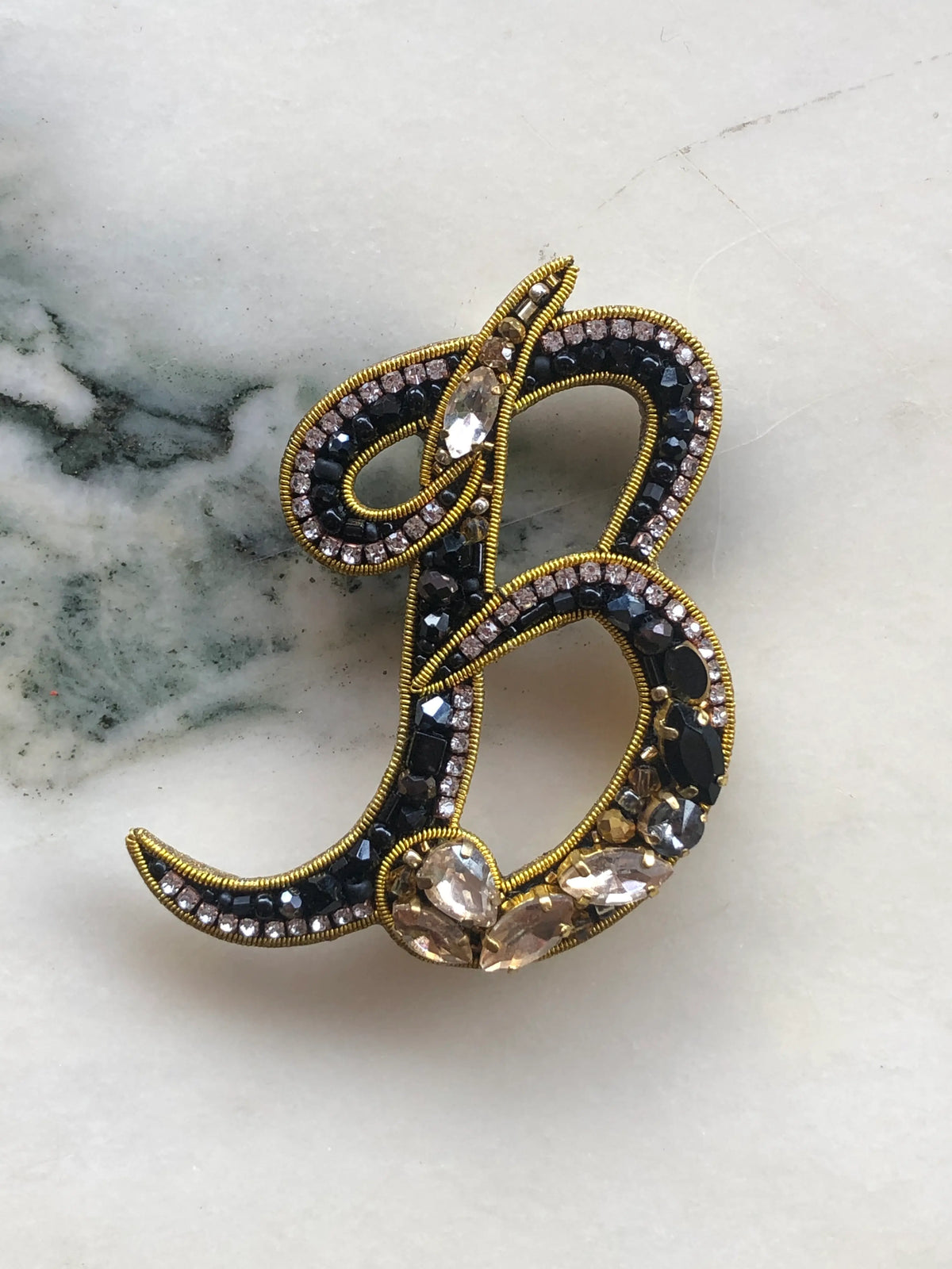 Broochella Men Initial B Brooch embellished with black beads, clear crystals and champagne stones with gold detailing, photographed on a white marble background with soft grey veining.