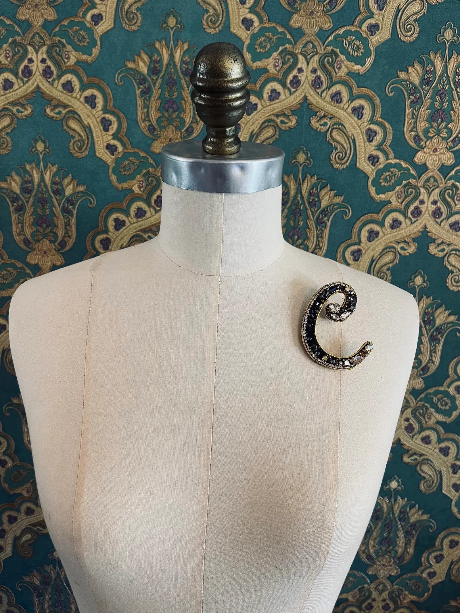 Broochella letter C hand beaded initial alphabet brooch in gold and black beads and sequins. Pictured on a mannequin. The background is green and gold damask wallpaper.