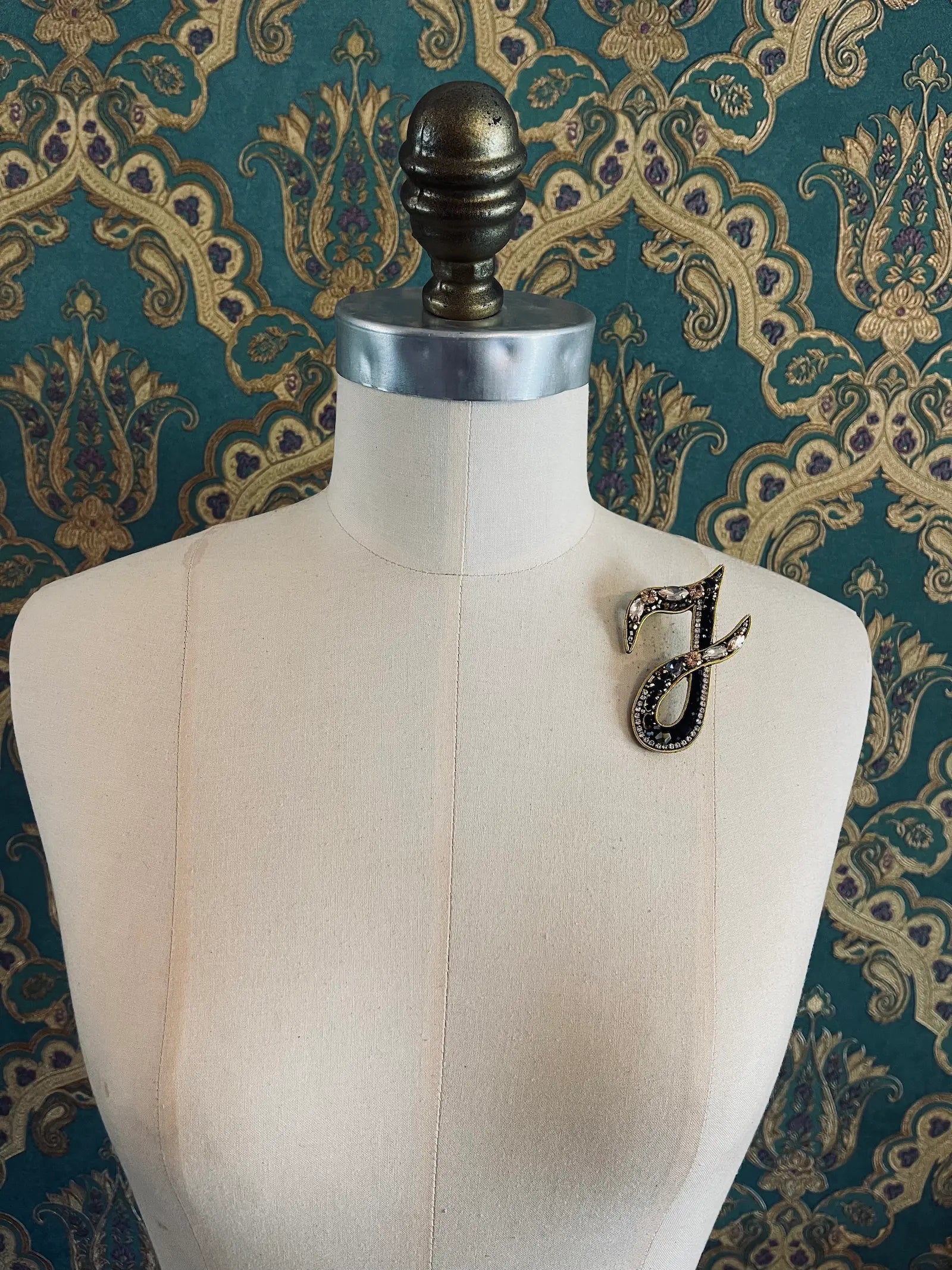 Broochella letter I hand beaded initial alphabet brooch in gold and black beads and sequins. Pictured on a mannequin. The background is green and gold damask wallpaper.