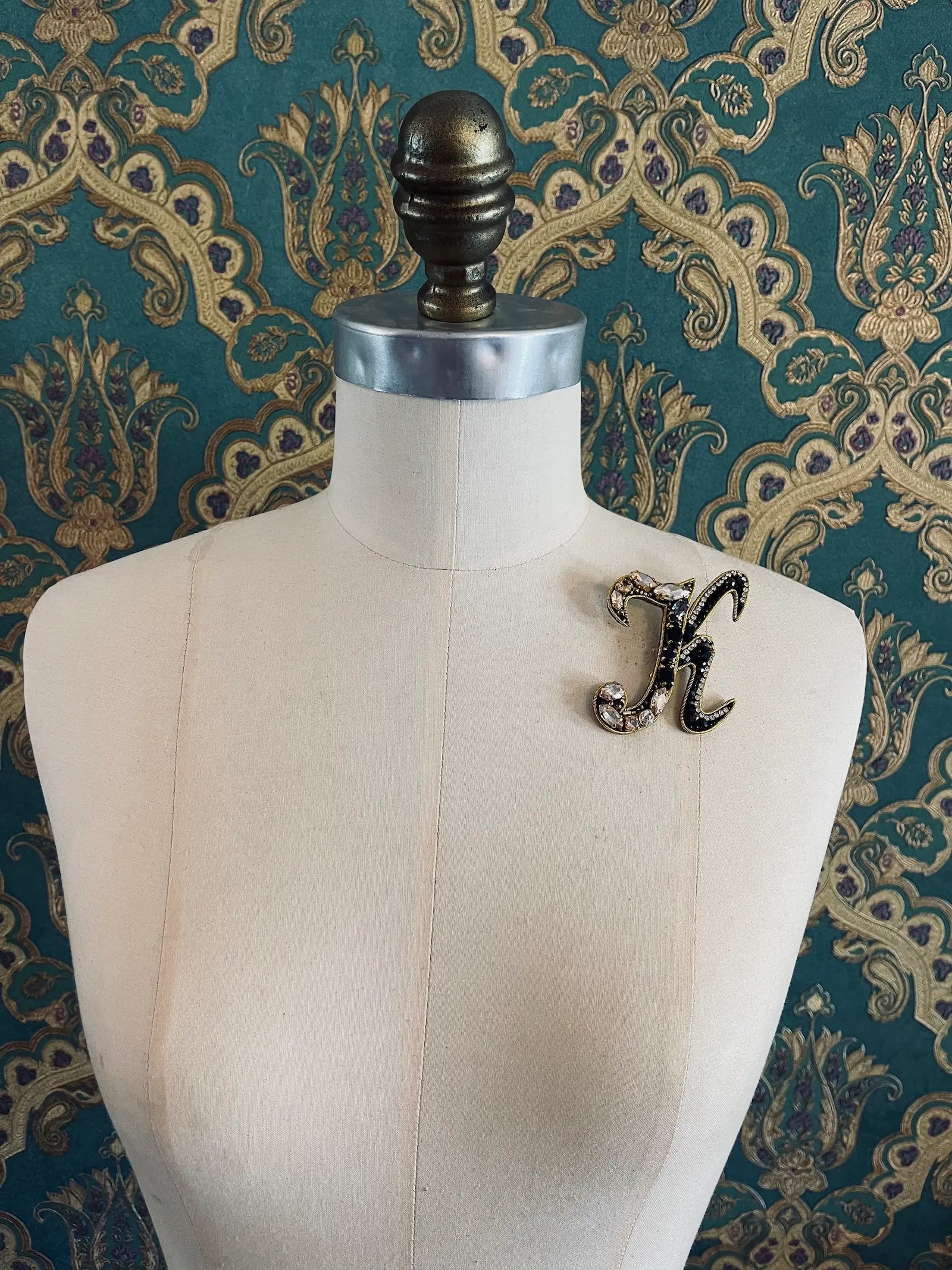 Broochella letter K hand beaded initial alphabet brooch in gold and black beads and sequins. Pictured on a mannequin. The background is green and gold damask wallpaper.